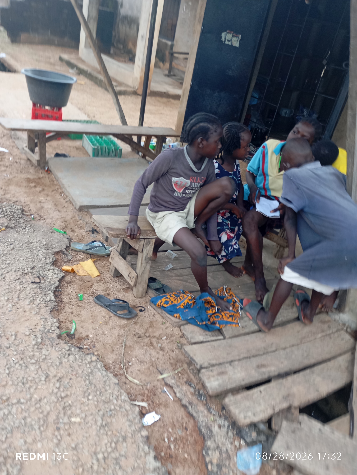 In Shendam, Nigeria, a group of children are gathered on and around a wooden bench outdoors. The scene appears to be a street or roadside setting with rough ground and a concrete slab. One child, wearing a grey shirt with "RUG" visible, sits on the bench. Another child in a patterned dress sits next to them. A third child, wearing a blue and yellow striped shirt, is seated, holding a white paper. A fourth child in a blue shirt is seen from behind, bending down. Several pairs of flip-flops are scattered on the ground. In the background, a large black basin sits on red crates, and a wooden structure with a metal grate is visible. The overall environment suggests a humble, possibly rural or semi-urban, community. The date stamp "08/28/2026 17:32" is present in the bottom right corner.