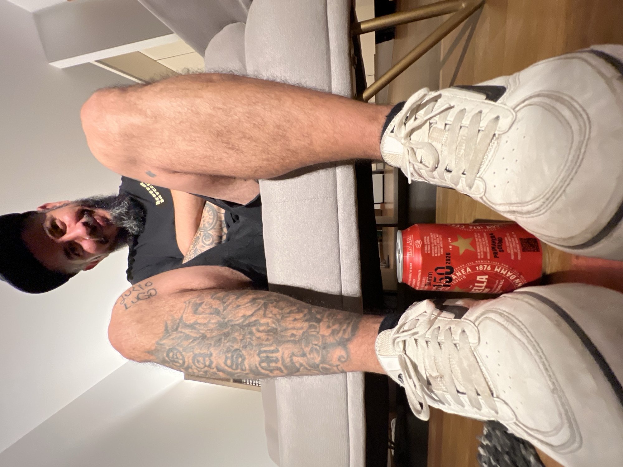 A man wearing a black t-shirt and cap is sitting on a gray sofa. His hairy legs are extended, revealing tattoos on his right leg. He is wearing white sneakers. Next to one of his sneakers is a red can of Damm beer, a beer from L'Hospitalet De Llobregat, Spain. The photo appears to be taken indoors. The floor is wooden.