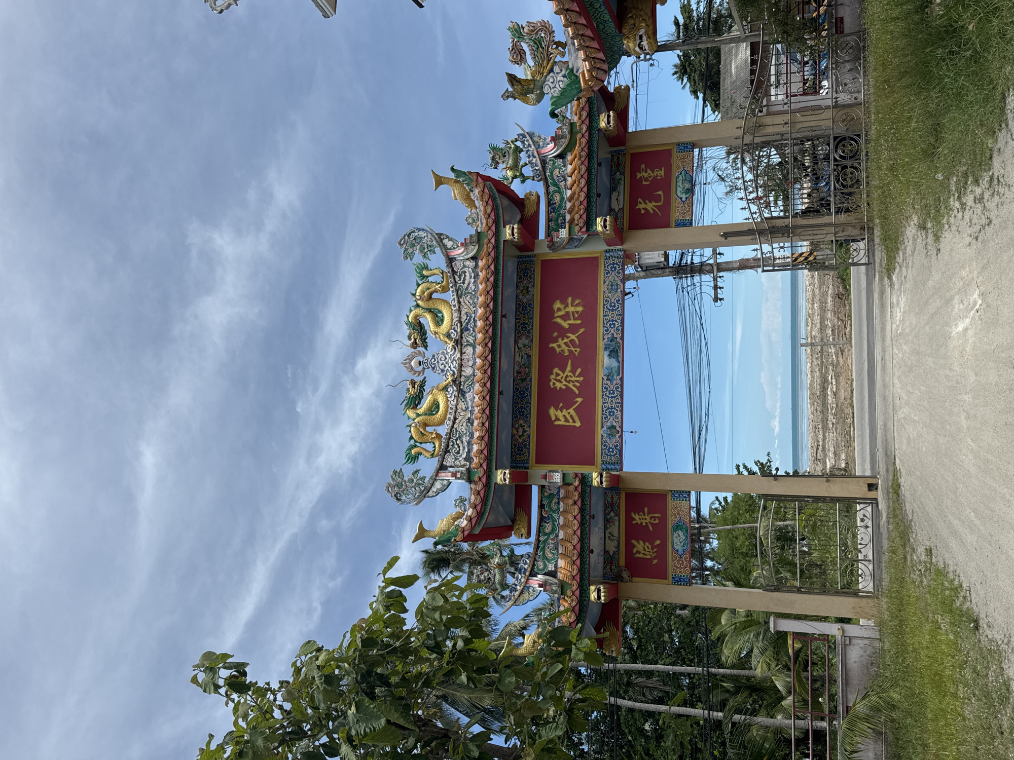 The image depicts a grand, ornate archway situated in Koh Phangan, Thailand, leading towards a beach. The archway is a traditional Chinese architectural style, featuring vibrant red and gold colors, intricate carvings, and dragon motifs atop its roof. A large red sign with golden Chinese characters is prominently displayed across the arch's peak, with smaller signs on either side.
Through the archway, a sandy beach stretches out to meet the calm, blue ocean under a partly cloudy sky. Palm trees and lush green foliage frame the scene on either side of the arch. A metal gate is partially visible at the entrance, suggesting a controlled access point to the area beyond. The overall impression is one of cultural richness and a picturesque coastal setting.