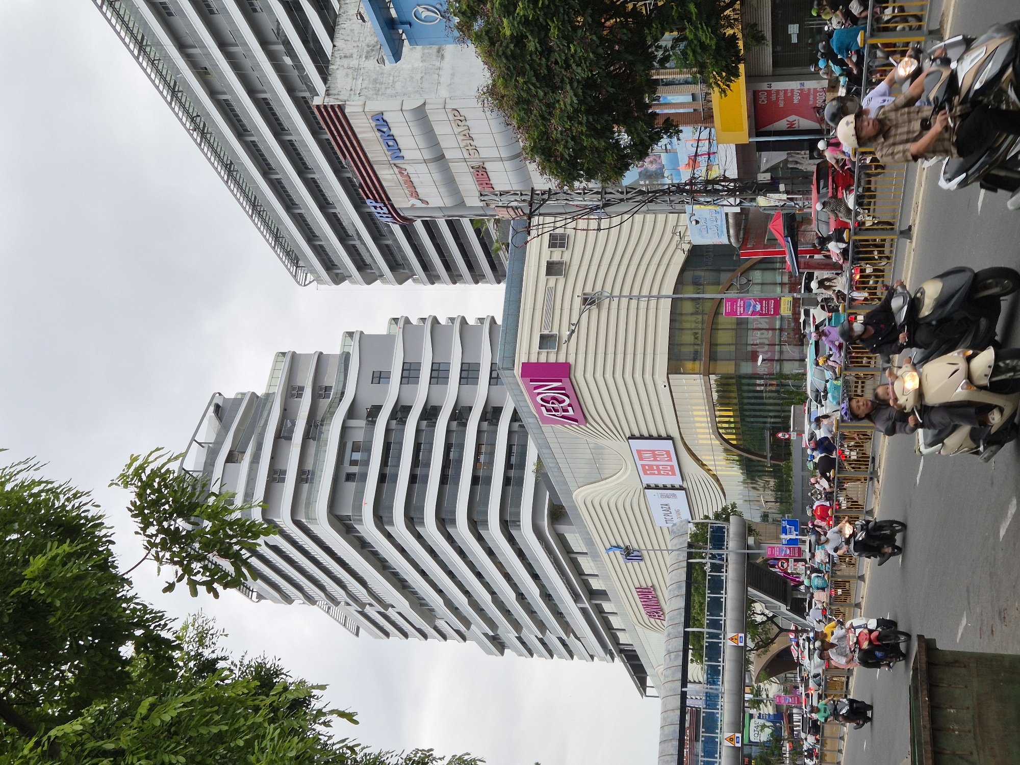 The image depicts a street scene in Da Nang, Vietnam, with a prominent modern building complex featuring a curved, multi-tiered facade. The complex appears to be a shopping mall or mixed-use development, with signs for "AEON MALL," "TTC PLAZA DA NANG," and "UNIQLO" visible. Adjacent to this, another tall building with a grid-like facade is visible, with signage for "NT&P," "SHARP," and "NOKIA." The street is busy with traffic, predominantly motorcycles, with many individuals riding them. Pedestrians are also present, with one woman carrying a child on her motorcycle in the foreground. The environment is urban, with trees lining the street and an overpass visible in the background. The sky is overcast, suggesting a cloudy day. The overall impression is one of a bustling city center in Vietnam.