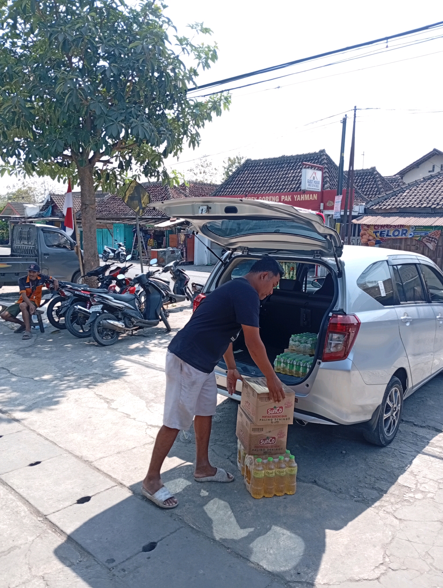 In Playen, Indonesia, a man is seen loading boxes of SunCo cooking oil into the open trunk of a silver car. The scene takes place on a paved street, with a tree providing shade to the left of the frame. Several motorcycles are parked nearby, and a grey truck is visible behind them. A person is seated on a small stool to the left, near the truck. In the background, buildings with tiled roofs are visible, suggesting a commercial or residential area. The sky is bright and clear, indicating a sunny day. The man is wearing a dark t-shirt and light-colored shorts, and he is placing two boxes of cooking oil on top of several stacks of bottled cooking oil. The overall impression is of a delivery or restocking activity.