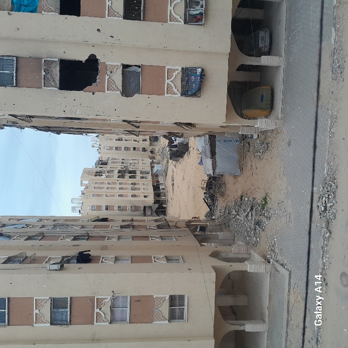The image depicts a scene of extensive structural damage within a residential area of Khan Yunis, Palestinian Territory. Two multi-story apartment buildings, each featuring a light beige facade with brown vertical panels and decorative white window frames, flank a central, open, sandy ground area.
The building on the left shows numerous damaged windows and missing panes, with some dark material or debris hanging from openings. The ground floor consists of open arches supported by pillars.
The building on the right exhibits more pronounced damage, including large, irregular holes in its facade and multiple shattered or missing windows, some covered with dark tarps or sheets. Debris is visible hanging from several openings. Underneath its ground-floor arches, a vehicle covered in yellow tarp is parked alongside a white sedan.
The central ground area between the buildings is unpaved, consisting primarily of sand and loose earth, heavily littered with concrete rubble, twisted metal, and assorted debris. A small, makeshift structure, covered with a white tarp, stands near the center, with another darker, less defined structure behind it. A lone, small figure, possibly a child, dressed in red, is partially visible near a pile of debris on the left side of the central area. In the background, additional multi-story residential buildings, similar in design and also appearing damaged, extend into the distance. The sky is overcast and uniformly grey. The bottom left corner of the image contains a "Galaxy A14" watermark.
