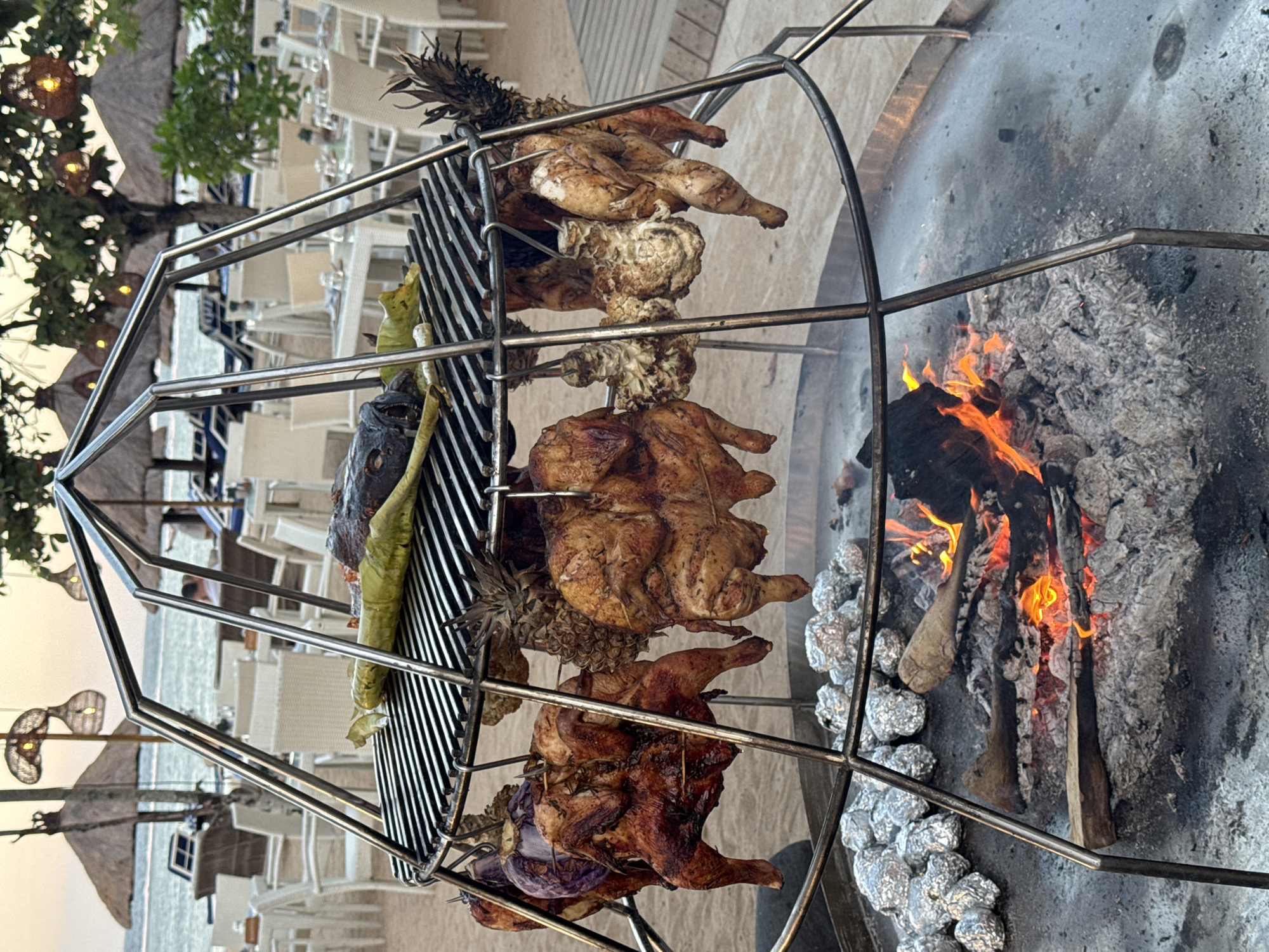 The image depicts an outdoor barbecue setup on a sandy beach, likely in Kuta Selatan, Indonesia, given the presence of palm trees, thatched umbrellas, and lounge chairs. In the foreground, a large metal rotisserie grill is suspended over a fire pit filled with burning logs and embers. Several whole chickens and an eggplant are skewered and rotating above the heat, glistening with juices. Some of the chickens appear to be golden brown and well-cooked, while others are still undergoing the grilling process. In the lower left of the image, a pile of potatoes wrapped in aluminum foil rests on the sand, presumably to be baked by the fire. The background shows a blurred view of tables and chairs set up for dining, with the ocean and horizon visible beyond. The lighting suggests it is either early morning or late afternoon, with the sky showing a warm, soft glow. There are no people clearly visible in the immediate vicinity of the grill.