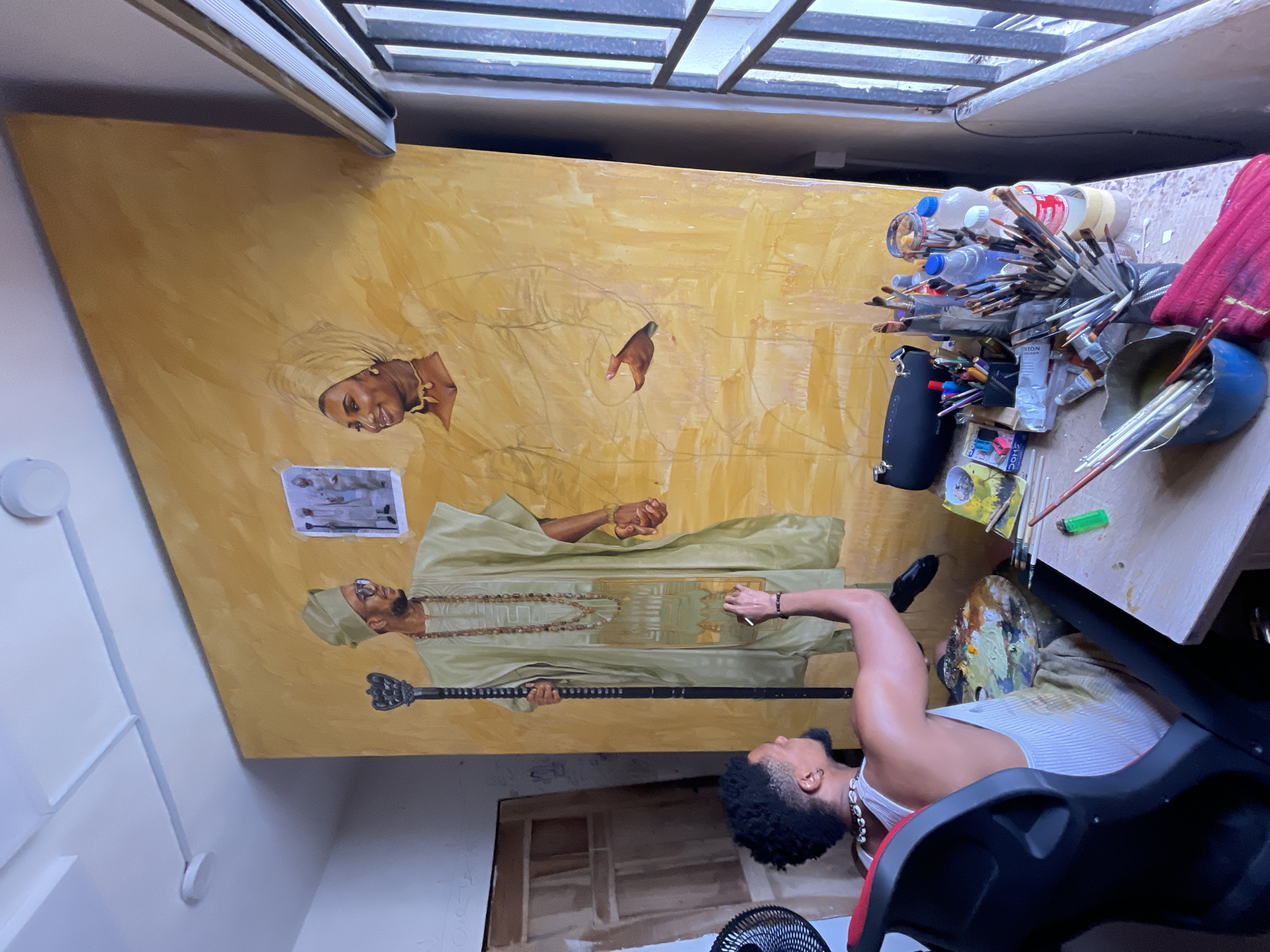 In Ado, Nigeria, an artist, identified as Oluwasegun "Segun" Ayo, is captured in his studio, actively engaged in painting a large portrait. The painting, displayed prominently, depicts a man and a woman in traditional attire against a warm, golden background. The man, on the left, is dressed in a pale green tunic and cap, adorned with beaded necklaces, and holds a staff. The woman, on the right, wears a matching pale green headwrap and garment, with a necklace. Above the figures, a small framed reference photograph is affixed to the canvas.
Segun, seated in a black and red ergonomic chair, is positioned to the left of the canvas. He is wearing a white tank top and appears to be working on the lower portion of the painting, holding a paintbrush in his right hand. A palette, laden with an assortment of paint colors, rests on his lap or in front of him.
The studio environment reveals a cluttered but organized workspace. A wooden table in the foreground is covered with art supplies, including various paintbrushes, tubes of paint, bottles of water, and a small green lighter. A portable speaker and other personal items are also visible on the table. Behind the artist, a partial view of another large artwork leans against the wall. The room is illuminated by natural light streaming in from a window on the right, which is covered by horizontal blinds and barred from the outside, suggesting security measures. The ceiling features simple white panels and visible lighting fixtures. The overall impression is that of a dedicated artist immersed in his creative process within his personal studio space.