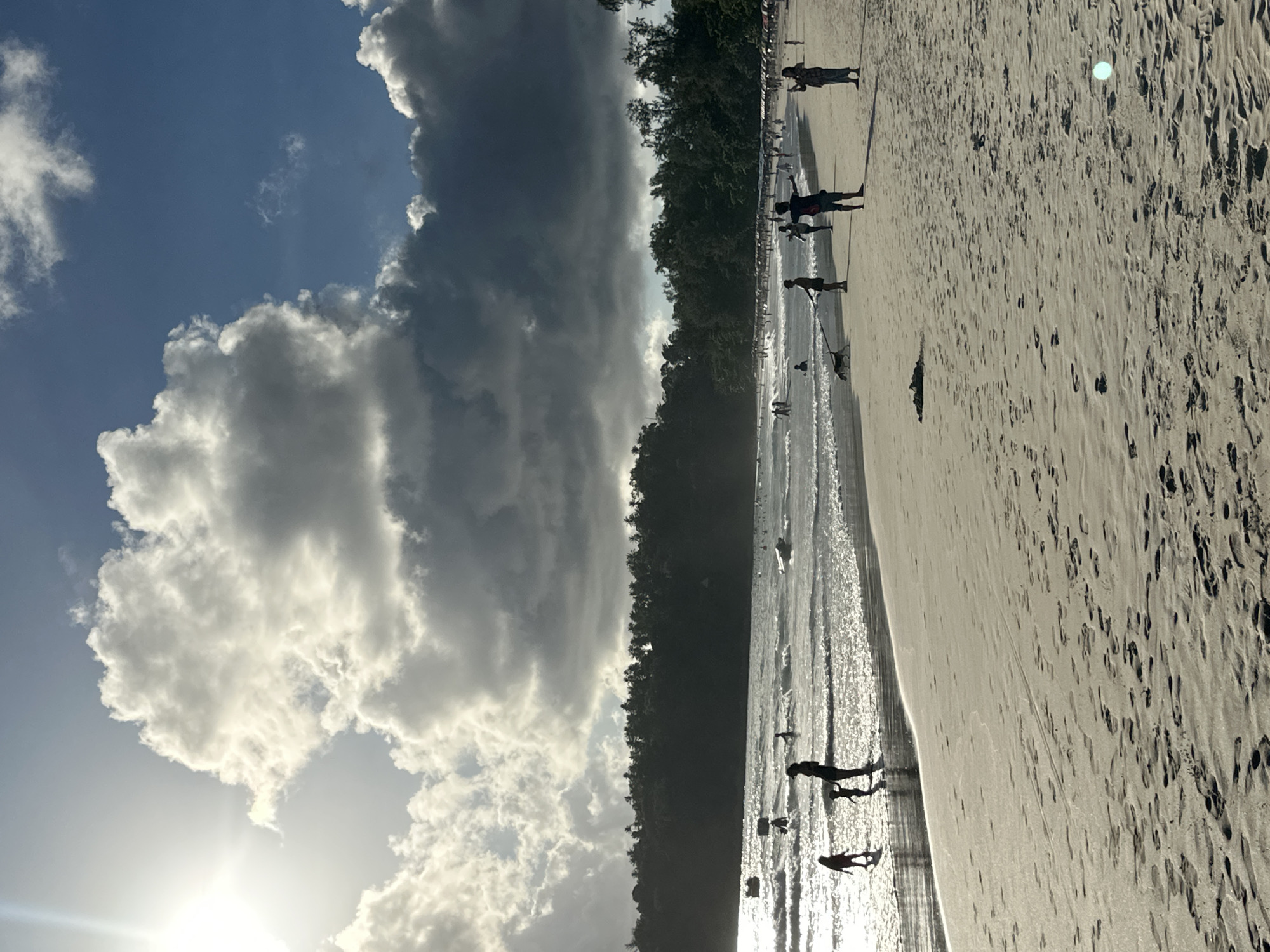 The image depicts a bustling beach scene at Karon Beach in Karon, Thailand. The sky is a dramatic mix of bright blue and dark, voluminous clouds, with the sun casting a strong glare from the upper left. The ocean is a vibrant turquoise, with moderate waves lapping against the shore. Numerous people are visible on the beach and in the water, engaged in various activities such as swimming, walking along the shoreline, and playing. The sand is light in color, marked with footprints and scattered debris. Lush, dark green foliage lines the background of the beach, suggesting a tropical environment. Several individuals are captured in silhouette against the bright sand and sea, including families with children and people walking dogs. The overall atmosphere appears lively and recreational, characteristic of a popular tourist destination.