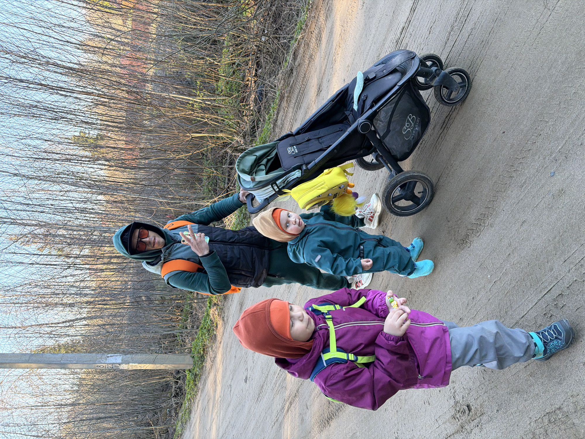 The image shows a man and two children outdoors, likely in a park setting. A stroller is parked nearby. The backdrop appears to be a wooded area with bare trees, suggesting it might be late fall or early spring. The ground is a dirt path. The older child is wearing a purple jacket, grey pants, and an orange hat. The younger child is wearing a green snowsuit and a hat. The man is in a green jacket and sunglasses. The stroller is black. Given the instructions, this image was taken in Chaikovsky, Russia.