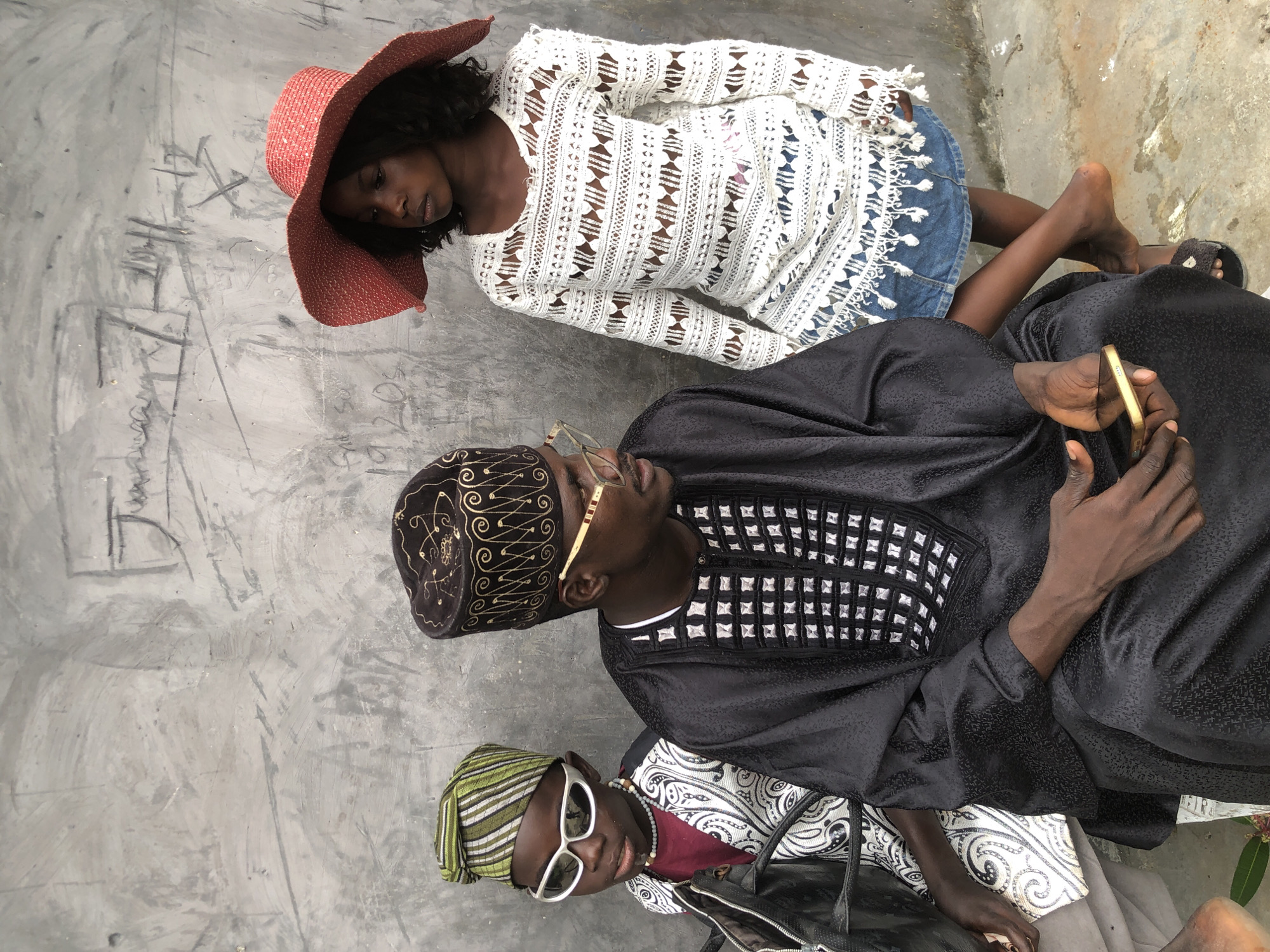 In Ado, Nigeria, three individuals are captured in a photograph against a gray concrete wall. On the left, a child with sunglasses and a striped green and white headwrap is dressed in a black garment with a white patterned vest. To their right, a man wearing a black cap with intricate gold embroidery and spectacles is adorned in a black robe. The robe features a squared pattern of small white embellishments around the neckline. The man holds a gold-colored smartphone in his right hand. On the far right, a young girl in a wide-brimmed red hat is seen from the chest down, wearing a white crocheted top over a denim skirt. The concrete wall behind them bears faint, scribbled markings. The ground is composed of concrete and dirt.