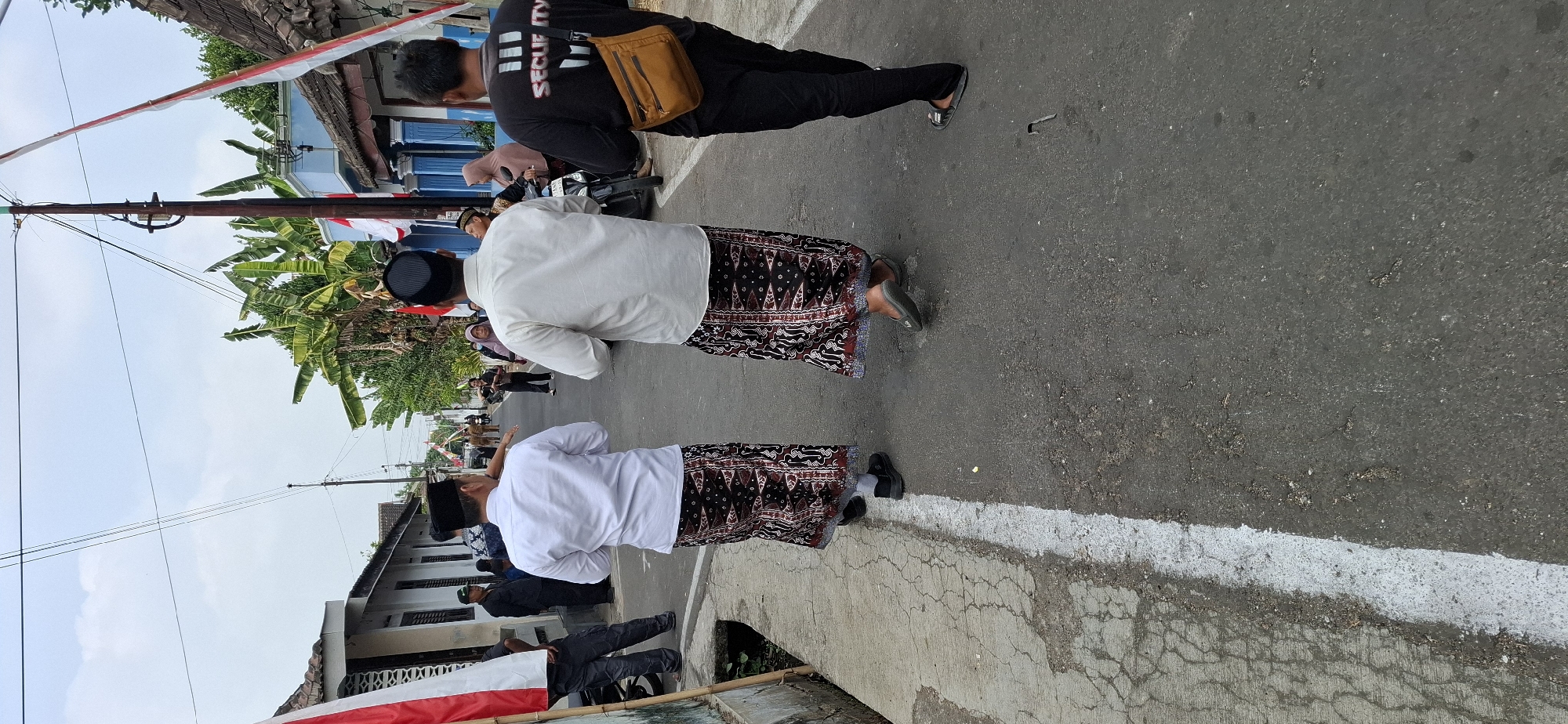 The image depicts a street scene in Gentan, Indonesia, with several individuals walking down a paved road. In the foreground, two men are seen from behind, both wearing white shirts and traditional sarongs with intricate patterns. They are walking in the same direction, towards the background. To the right of these two men, a third person, also seen from behind, is wearing a black t-shirt with "SECURITY" printed on the back and carrying a tan crossbody bag. The road has a white line marking its edge on the left side, adjacent to a concrete walkway. The environment includes buildings on either side of the street, with some greenery visible, including banana trees. There are also Indonesian flags displayed, suggesting a possible cultural event or celebration. Other people are visible in the middle ground and background, some of whom appear to be onlookers. The sky is overcast, indicating a daytime scene.