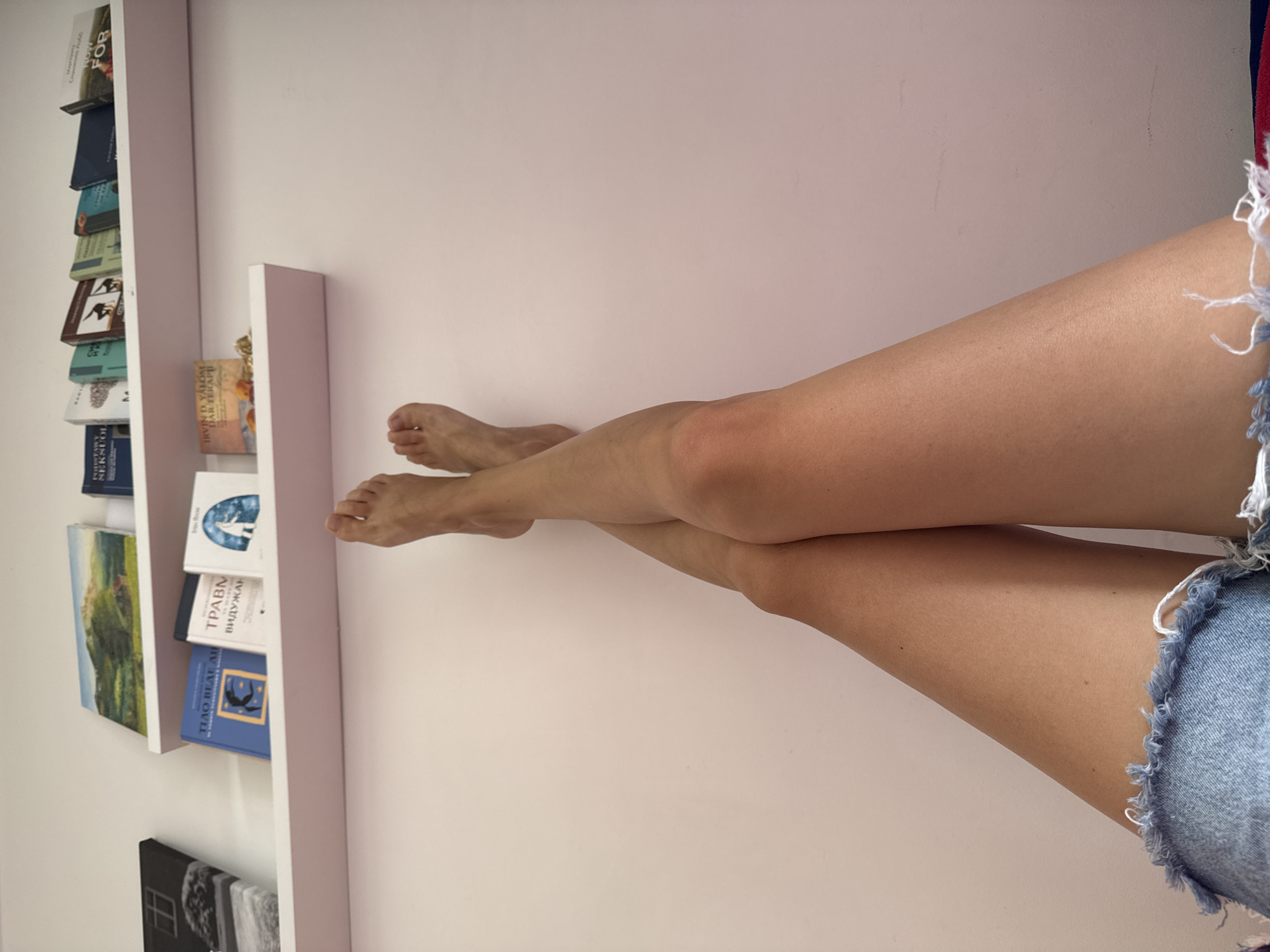 The image presents a low-angle view looking upwards at a person's legs and two wall-mounted shelves against a light-colored wall. The scene is an interior, located in the city of Unknown, Unknown.
In the foreground, a person's bare legs are crossed at the ankles, extending upwards from the bottom of the frame. The feet are visible at the top of the legs, pointing towards the upper part of the image. The skin exhibits a light tan. A portion of distressed blue denim fabric with frayed edges, indicative of shorts, is visible at the very bottom edge of the frame.
Above the legs, two identical light-colored, possibly pink or white, floating shelves are mounted on a plain, light pink wall.
The lower shelf holds three books standing upright and one smaller book lying flat in front of them. From left to right: a blue book with a title partially visible as "ТЬАО БЕЛ АН" (Tjao Bel An) and a silhouette of a dancing figure; a white book titled "ТРАВМ" (Travm) in Cyrillic script; and another white book featuring an illustration of a robed figure within an archway. The book lying flat is an orange-covered volume with text "IRVIN D. YALOM ДАР ТЕРАПИ" (Irvin D. Yalom The Gift of Therapy).
The upper shelf contains several books and two distinct pieces of artwork. On the far left, a black and white image, possibly a window or architectural detail, is mounted on a canvas or frame. To its right, a landscape painting or print depicting green hills and a small building is visible. A row of books stands upright on this shelf, some with partially visible Cyrillic titles such as "НЕКСУОЙ" (Neksuoi) on a blue book and "HOW FOR" (likely "How to...") on a small white book near the right. The books display various cover colors including blue, white, green, and brown.
The overall lighting is bright and even.