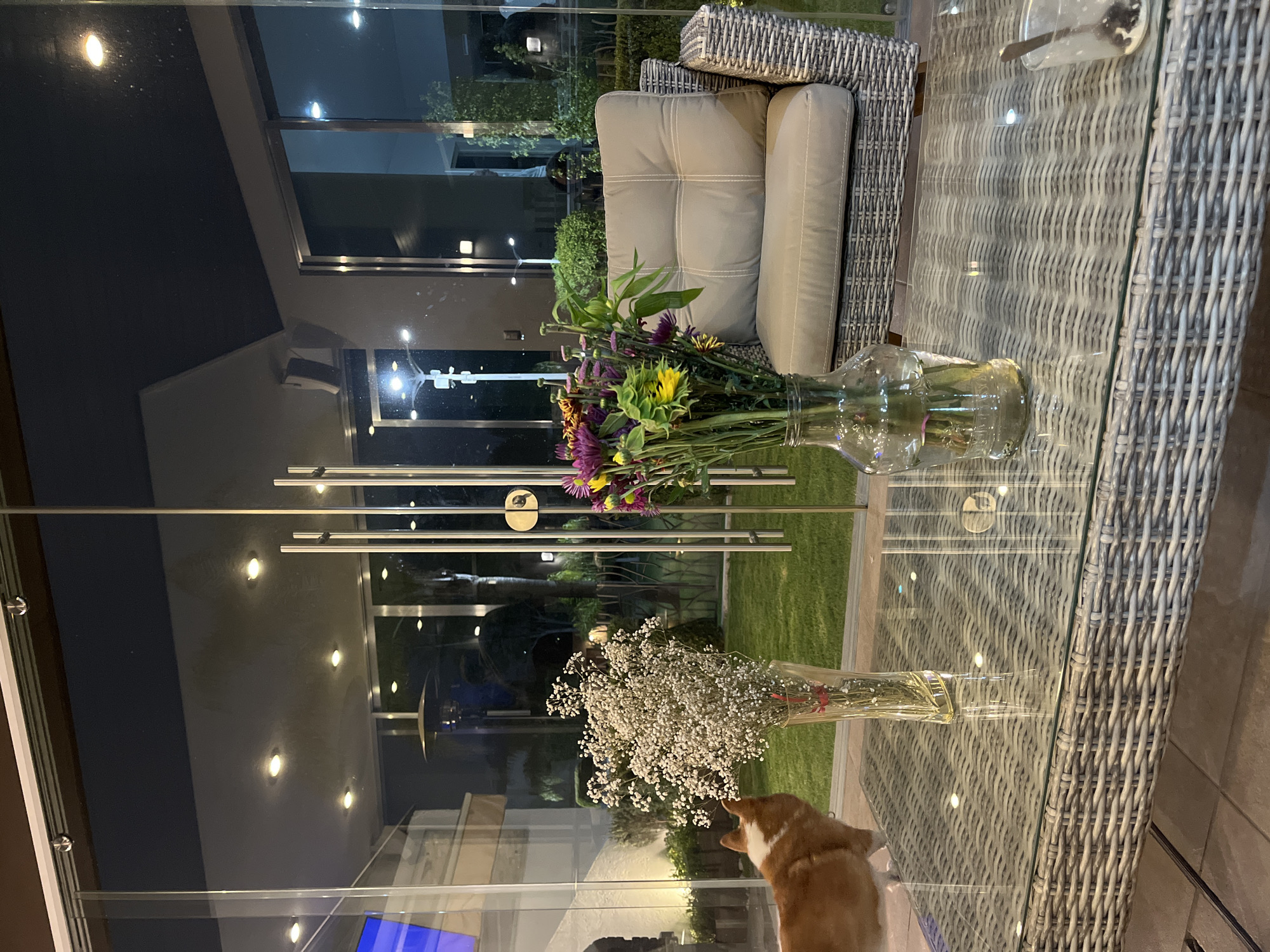 A photo taken at night in San Bernardino Tlaxcalancingo, Mexico shows a glass-topped wicker table with two vases of flowers. One vase holds white baby's breath flowers, while the other contains a colorful mix of flowers including yellow sunflowers and purple blooms. Part of a light beige wicker sofa with cushions is visible behind the table. Outside, a house and yard with green grass can be seen through the glass, illuminated by outdoor lighting. The side of a red and white dog is partially visible at the edge of the table.