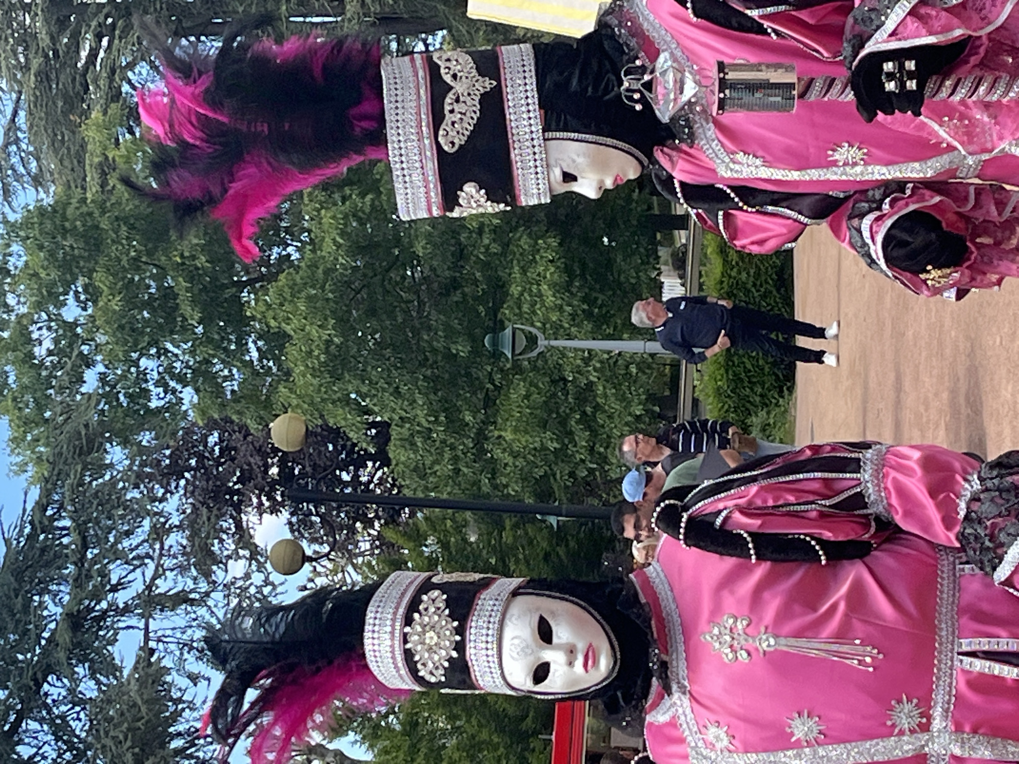 Two people dressed in elaborate Venetian costumes stand in a park-like setting in Saint-Galmier, France. The costumes are predominantly pink and black, heavily adorned with silver trim, jewels, and ornate headpieces with black and pink feathers. Both figures wear white masks. In the background, other people can be seen walking on a paved path, along with trees and street lamps. The scene is captured outdoors during the daytime.