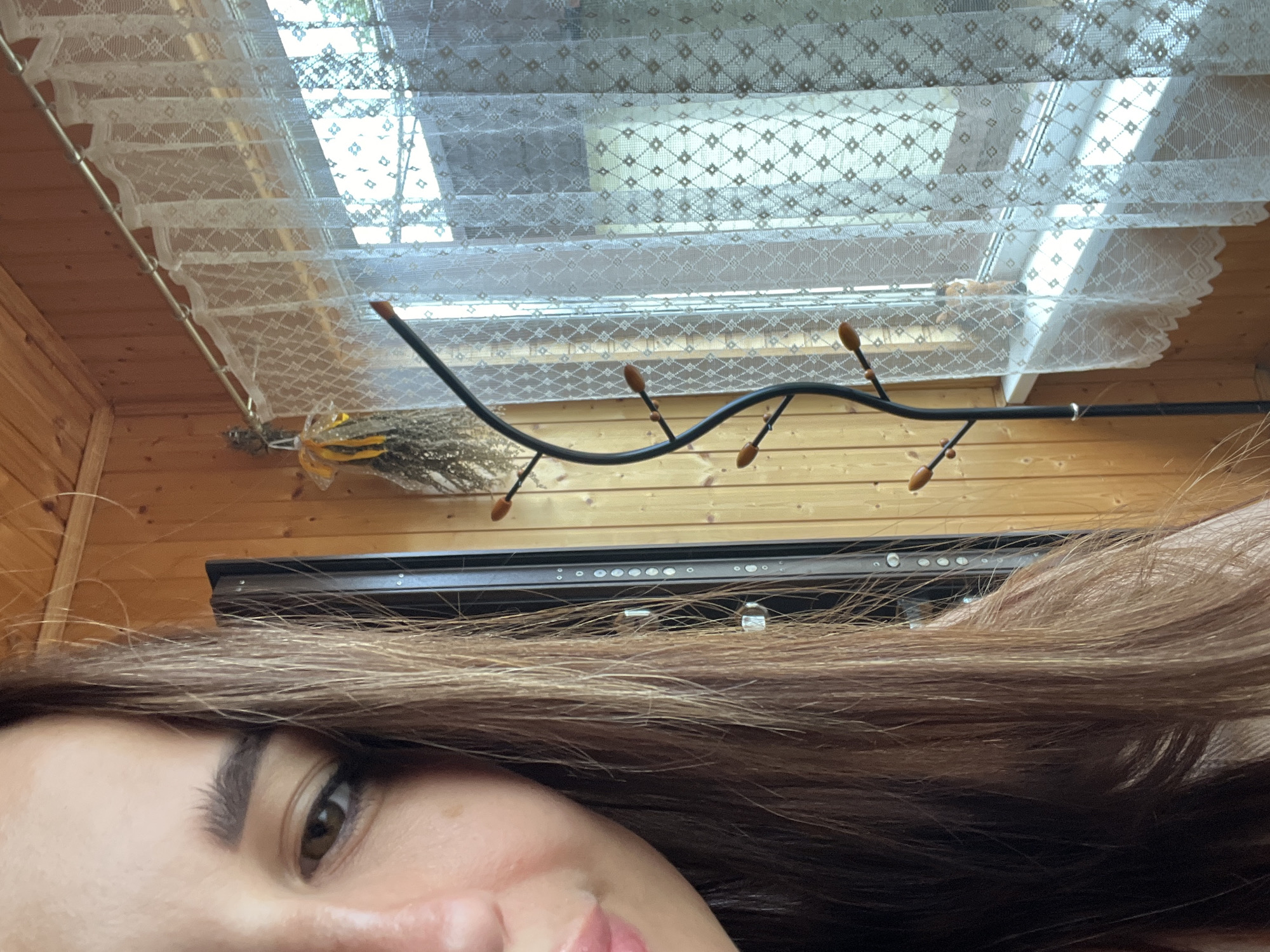 The image shows a close-up of a woman's face, with her left eye, eyebrow, and part of her mouth visible. She has long, brown hair that is partially obscuring her face. The background is a room with wooden walls. To the right of the woman, there is a window with a white lace curtain. A black coat rack with wooden hooks is positioned in front of the curtain. A dried floral arrangement hangs on the wall to the left of the curtain. The scene appears to be indoors, possibly in a rustic or country-style setting.