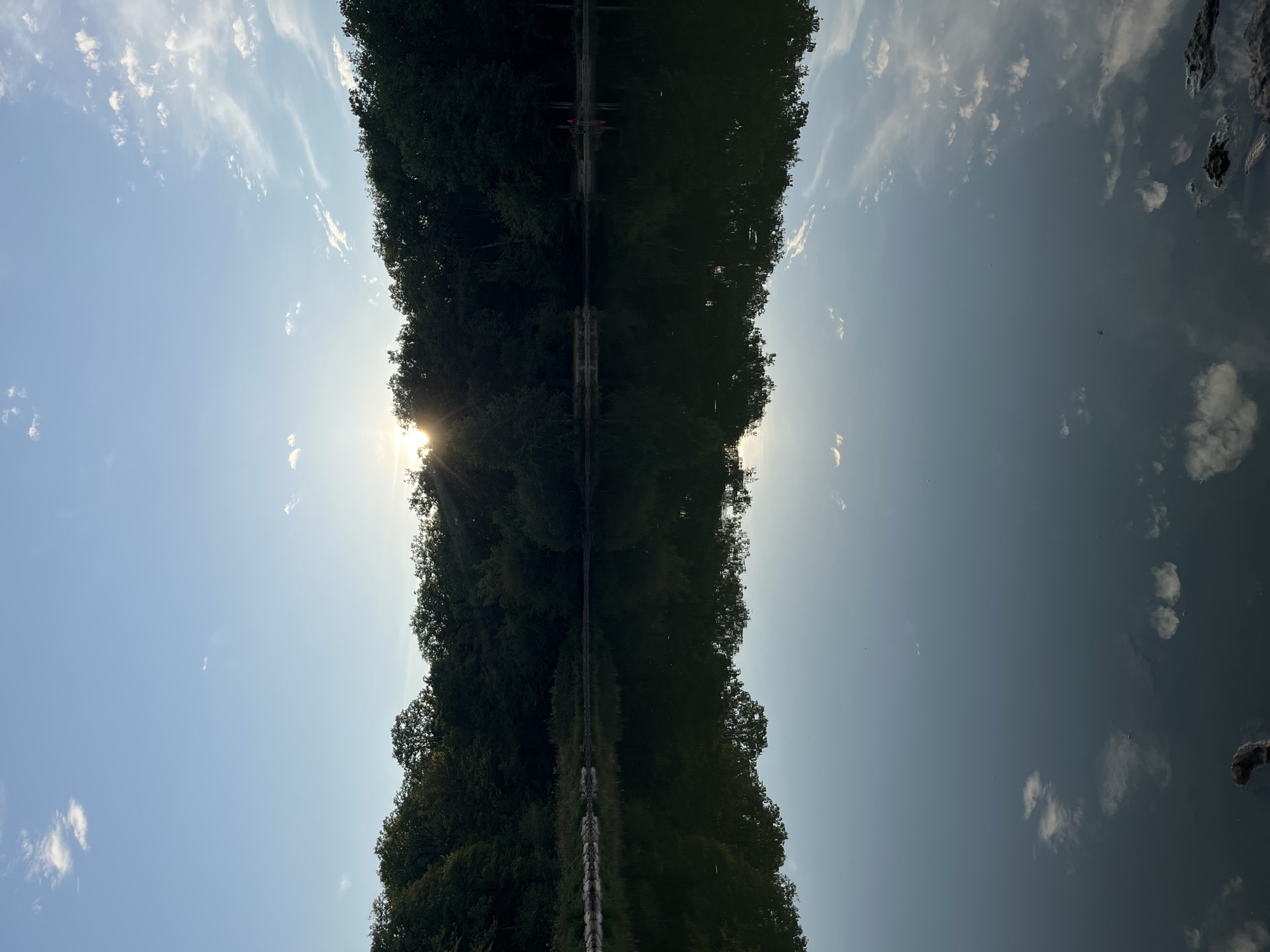The image depicts a tranquil lake at dusk, situated in Sonchamp, France. The water's surface acts as a mirror, reflecting the surrounding dense forest and the pale blue sky dotted with wispy clouds. The sun, low on the horizon, peeks through the trees, casting a warm glow and creating a silhouette of the forest against the sky. The water is calm, with only slight ripples disturbing the perfect reflection. The foreground shows a few scattered rocks and a piece of wood on the bank, hinting at a natural, undeveloped shoreline. In the distance, two figures, possibly on the far bank, are barely discernible against the dark trees, adding a subtle human element to the serene natural landscape.