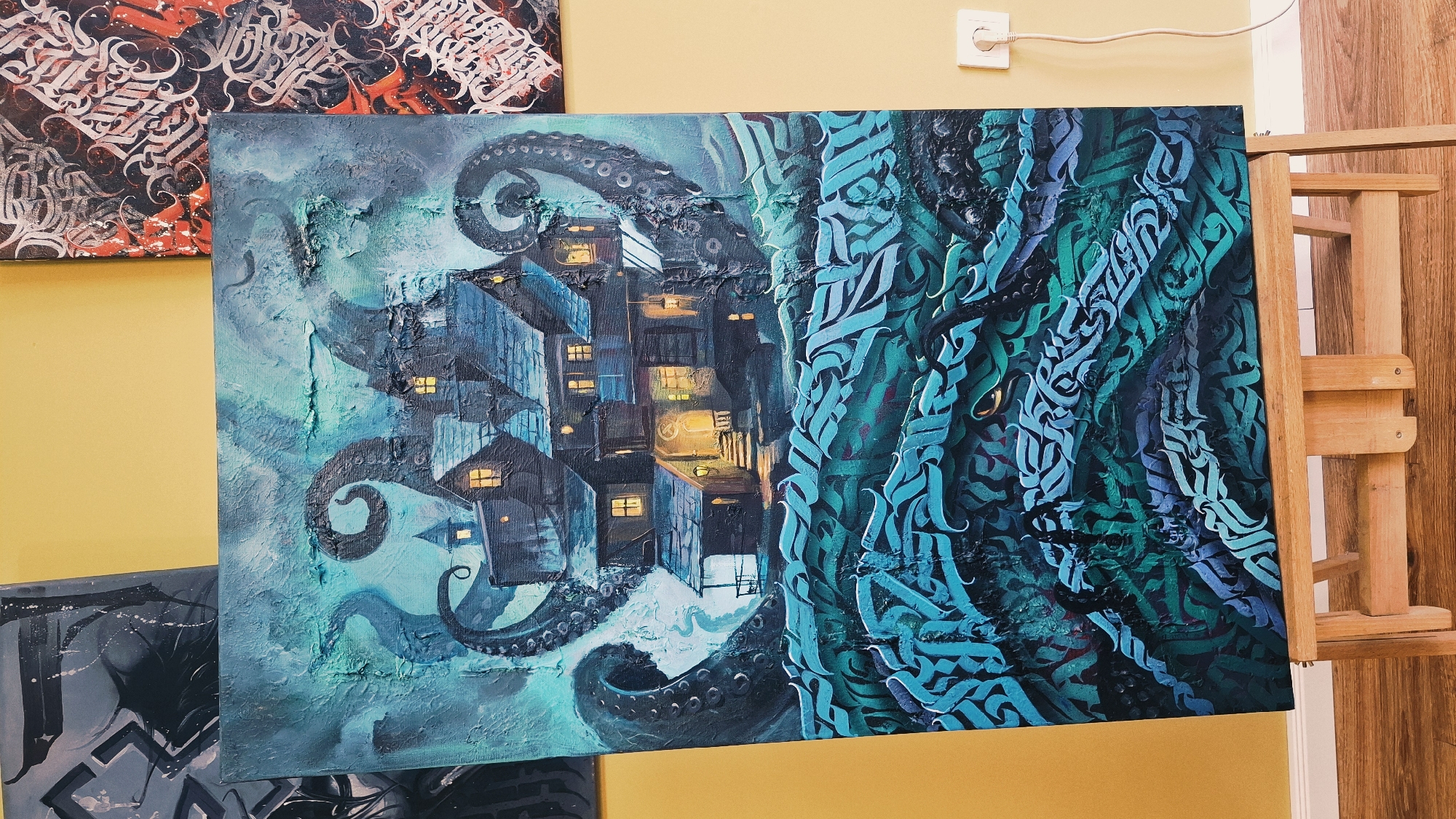 The photograph depicts a painting of a dark, haunted-looking house being attacked by a kraken. The house is set on a stormy sea, with tentacles of the creature wrapping around it. The painting is displayed on a wooden easel in what appears to be an indoor art exhibition or studio. Two other paintings are visible in the background, one on the left and one on the right, both with abstract designs. A yellow wall and a wooden floor are also present in the scene. The painting itself is textured and uses a palette of dark blues, greens, and hints of yellow light emanating from the house's windows. The bottom portion of the painting features abstract, wave-like forms with calligraphic elements in shades of turquoise and dark blue. A white electrical outlet with a white cord is visible on the yellow wall behind the easel. The image does not contain any specific details that would allow for identification of its location in Plovdiv, Bulgaria.