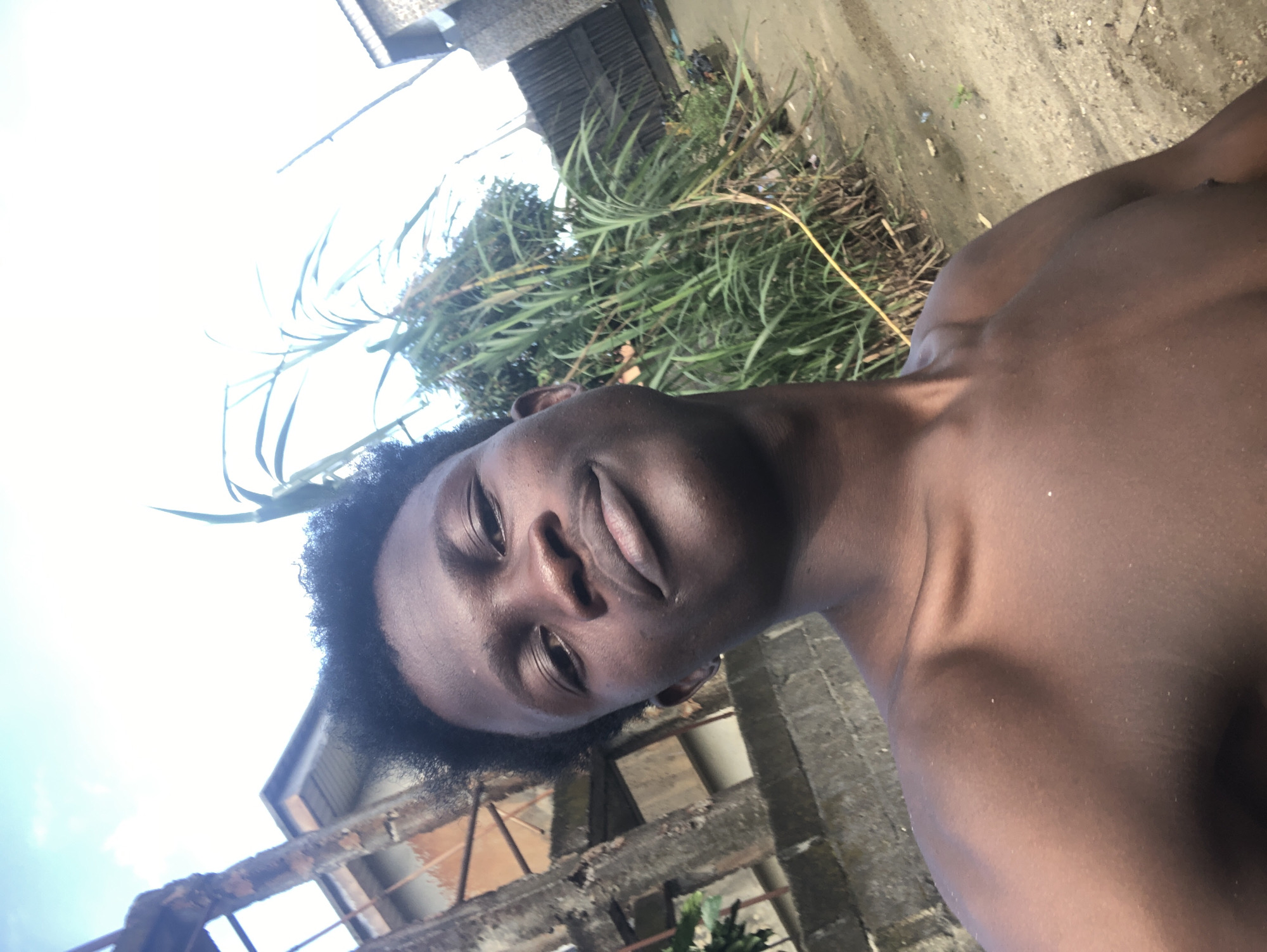 A young Black male, shirtless, takes a selfie with his chest and face filling the foreground. He is outdoors, with a bright sky and clouds visible in the upper portion of the image. Behind him, tall green reeds or grass grow, with some foliage and trees interspersed. To the left of the reeds, a weathered, multi-story building with visible rebar and concrete structures stands, appearing to be under construction or in disrepair. A gray concrete wall runs horizontally across the mid-ground. The ground beneath the reeds and building appears to be dirt or sand. To the far right, a corrugated metal fence is visible, suggesting a boundary. The lighting suggests it is daytime, likely late afternoon or early morning due to the angle of the light casting subtle shadows on his skin. The overall environment appears to be an urban or semi-urban setting in Ado, Nigeria, possibly a residential area or a lot adjacent to buildings, with evidence of ongoing construction or a more dilapidated state of infrastructure.