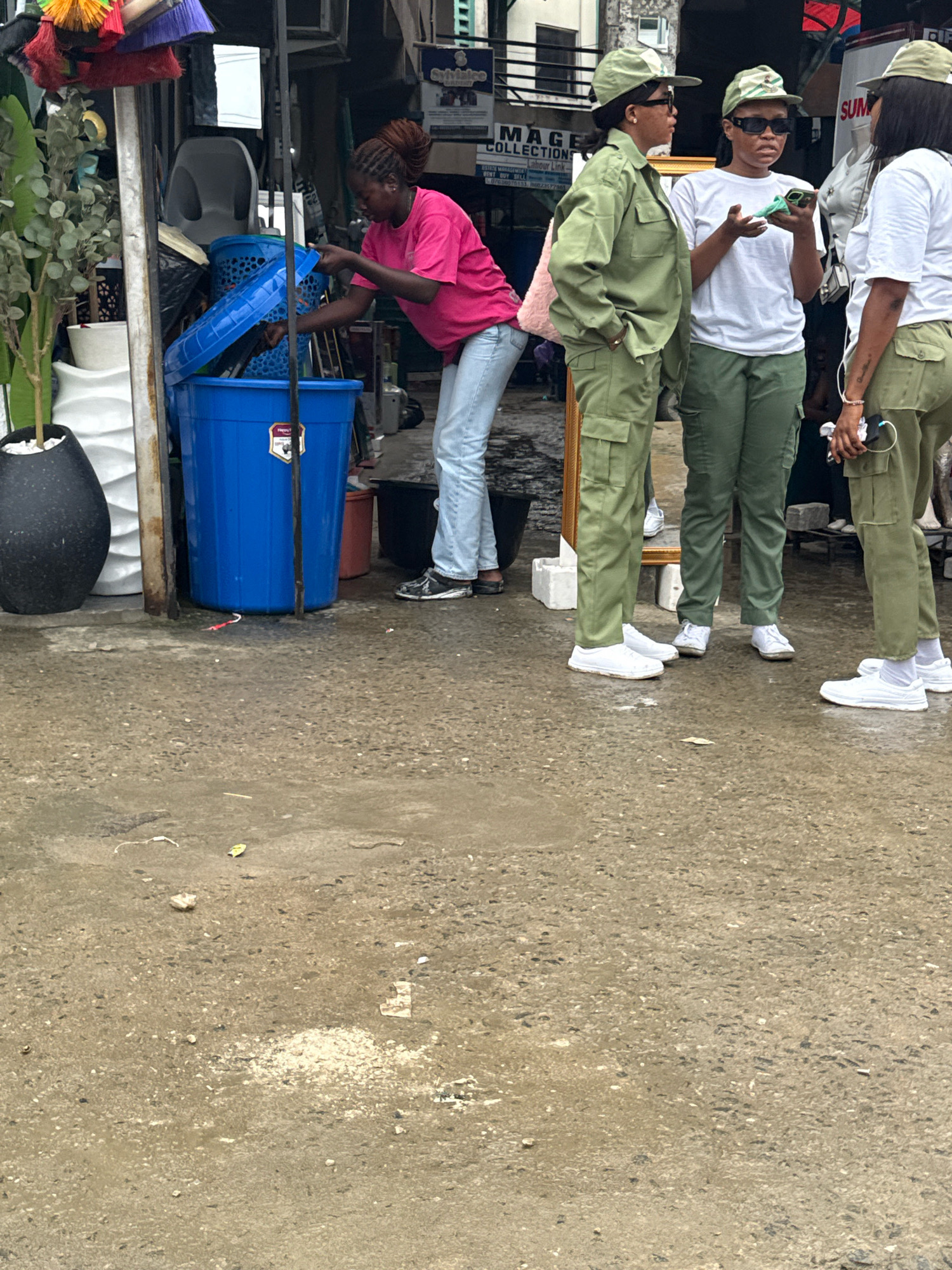 The image depicts an outdoor scene in Lekki, Nigeria, with a focus on a group of people and their surroundings. The ground is wet and appears to be paved or concrete, with puddles and debris scattered across its surface, suggesting recent rain.
In the foreground, a young woman wearing a pink t-shirt and light blue jeans is interacting with a large blue plastic bin. She is leaning over the bin, her right hand reaching into it as she holds open a blue lid. Next to her, to her right, are three individuals dressed in military-style green uniforms and matching caps. They are standing in a group, and the two further to the right are holding mobile phones. One of them is using her phone, while the other appears to be looking at hers. The person in the middle of the uniformed group is looking towards the woman with the bin.
Behind the individuals, the scene reveals a cluttered stall or shop. Visible items include a black plastic bin, a red bucket, a tall black vase containing a potted plant, and various other objects that suggest a market or retail environment. There are also brushes and cleaning tools hanging from a structure above the stall. The overall atmosphere suggests a bustling area, possibly a street market, in Lekki, Nigeria.