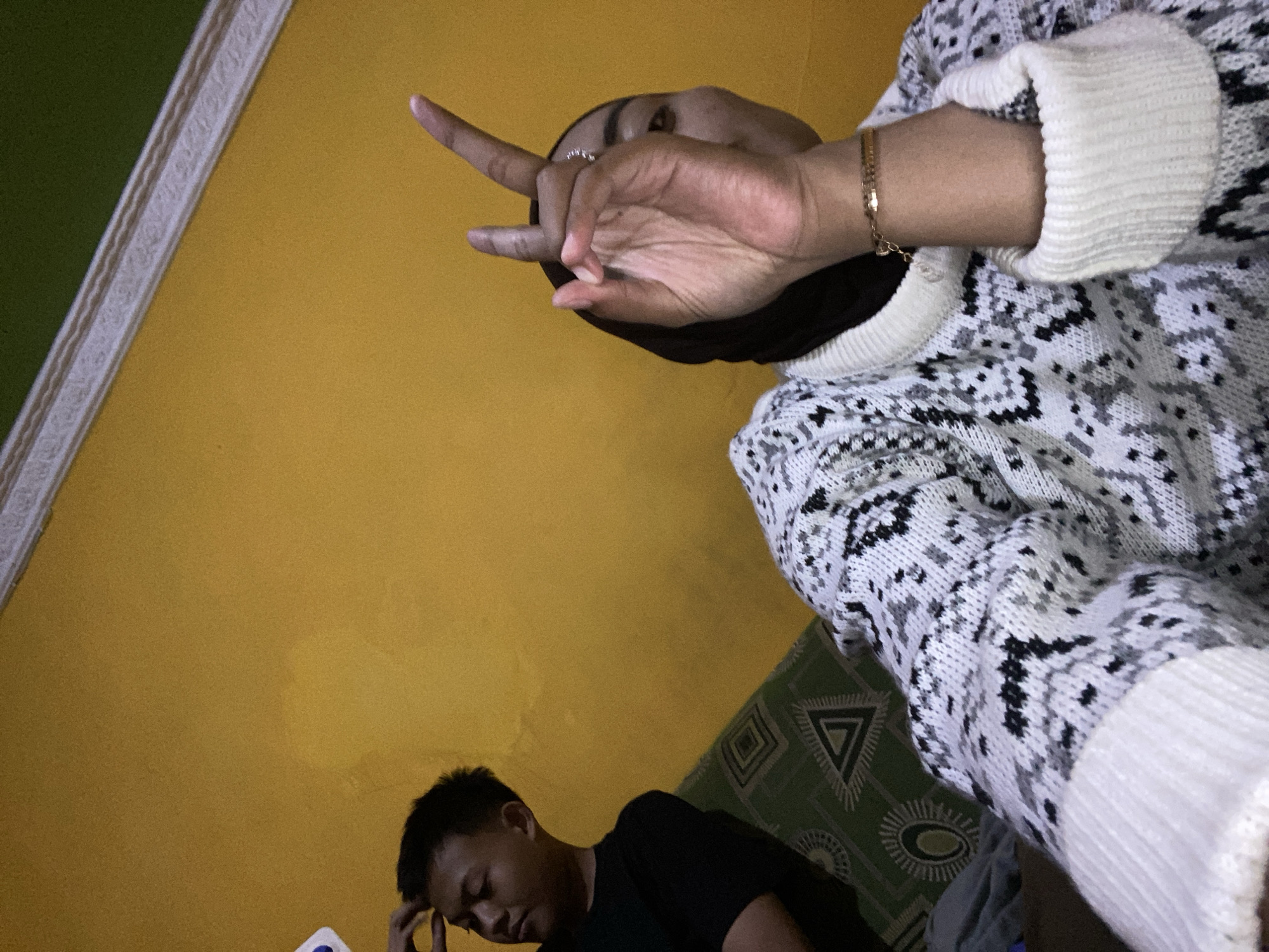 In Kecamatan Pati, Indonesia, a young man in a black t-shirt is seated on a patterned sofa, his hand resting on his forehead. Beside him, a person wearing a black hijab and a patterned sweater is holding up two fingers in a peace sign, their face partially obscured. The room features a bright yellow wall and a white ceiling with decorative molding. The sofa has a green and white geometric pattern.