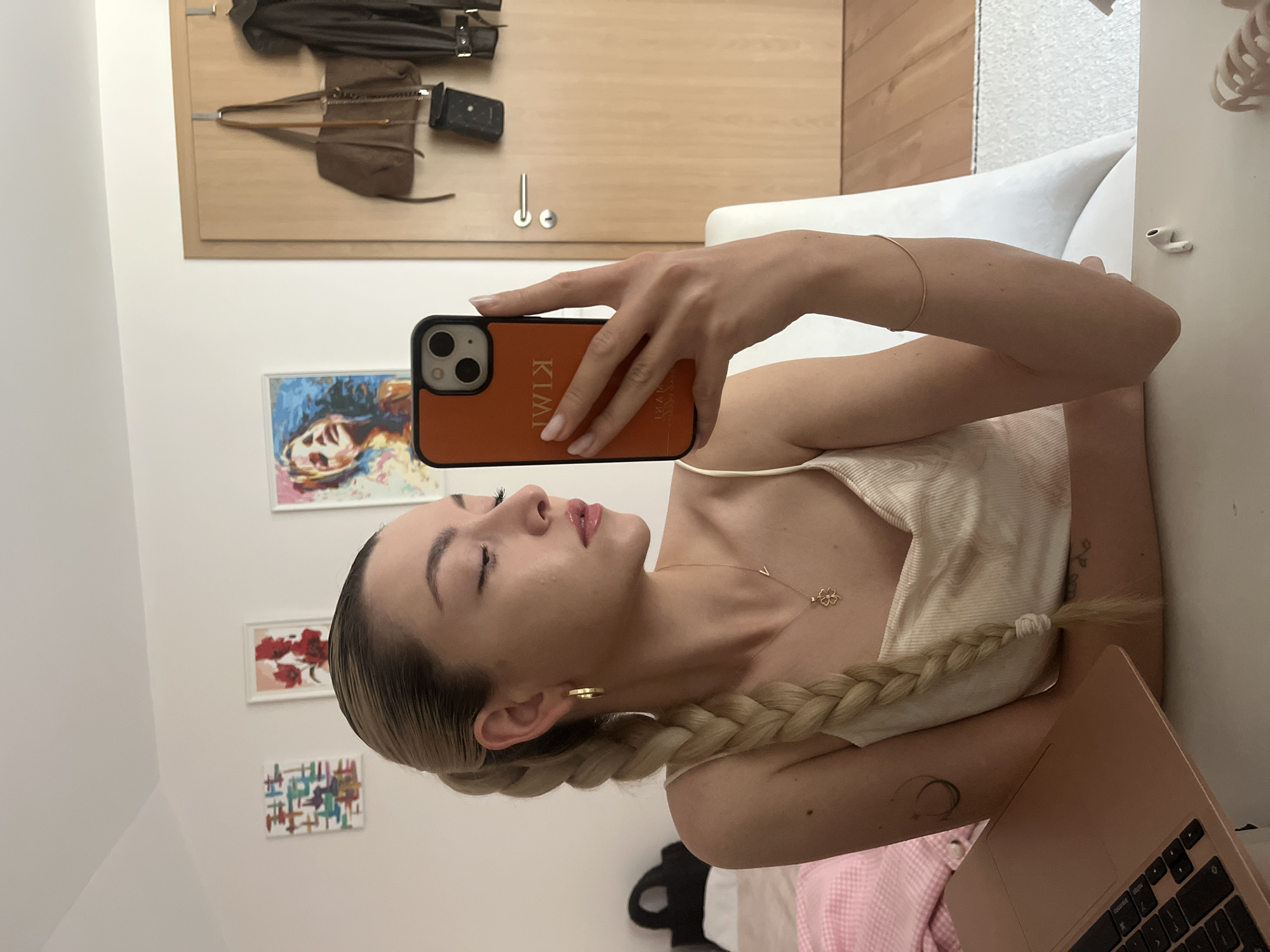 A young woman is taking a selfie with a cell phone that has a case on it with the name "KIMI" vertically on the back. She is wearing a light-colored tank top and gold jewelry, including a necklace with a clover pendant and hoop earrings. Her blonde hair is styled in a long braid.
She is indoors, possibly in a room in the city of Unknown, Unknown, with paintings on the wall behind her. To the right, there's a white couch and part of a laptop is visible, suggesting she may be working or relaxing. Behind her are an arrangement of coats and bags hanging on the wall. The overall lighting appears to be soft and natural.