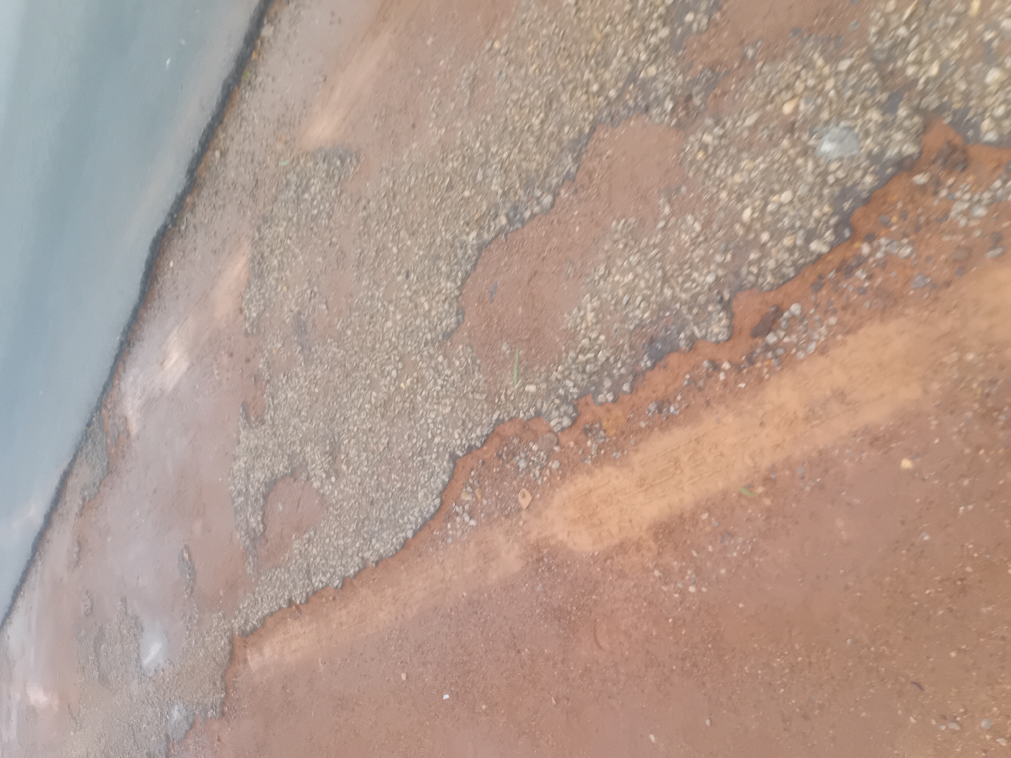 The image depicts a section of a damaged road in Jalingo, Nigeria. The road surface is heavily deteriorated, with large patches of exposed gravel and dirt. Some areas appear wet, indicating recent rainfall or standing water. The road runs diagonally from the bottom left to the top right of the frame, with a body of water, possibly a river or a large puddle, bordering the upper right edge. The overall impression is one of neglect and disrepair. No people or vehicles are visible in the image.