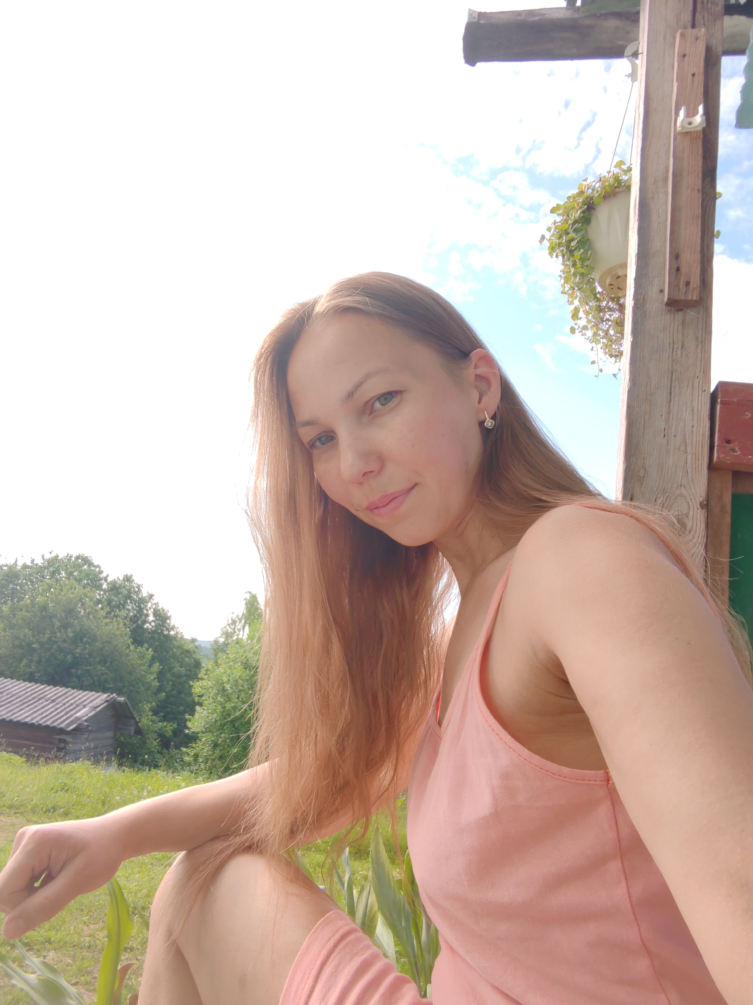 A woman with long, light brown hair and a pink tank top is sitting outdoors in front of a wooden structure with a hanging plant. She is looking at the camera with a slight smile. In the background, there are green trees, a small wooden building with a gray roof, and a bright blue sky with white clouds. The location is identified as Chukhlem, Russia. The image appears to be a selfie taken on a sunny day.
