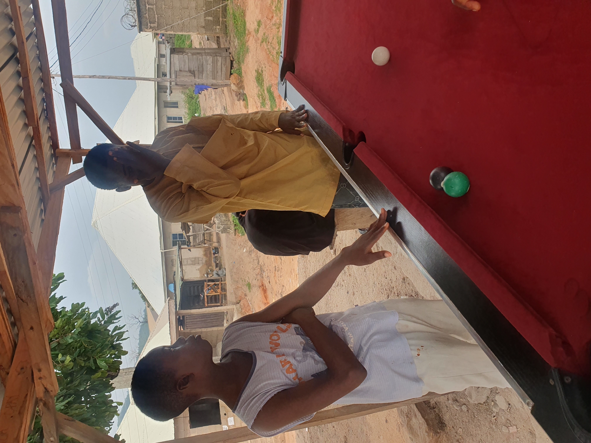 The image depicts two individuals standing near a red billiard table under a rustic wooden shelter in Lokoja, Nigeria. On the left, a young person wearing a white sleeveless shirt and light-colored pants stands with their arms crossed. To the right, an adult, partly obscured, is covering their face with their hand and wearing a mustard-yellow long-sleeved garment. The billiard table has several balls visible: a white ball in the upper right quadrant, and a green and a dark colored ball closer to the left side of the table. In the background, there are residential structures with white, sloped roofs and other buildings, some appearing under construction or in disrepair. The ground is dirt and gravel. The overall setting suggests an outdoor leisure area in a semi-urban or rural environment within Lokoja.