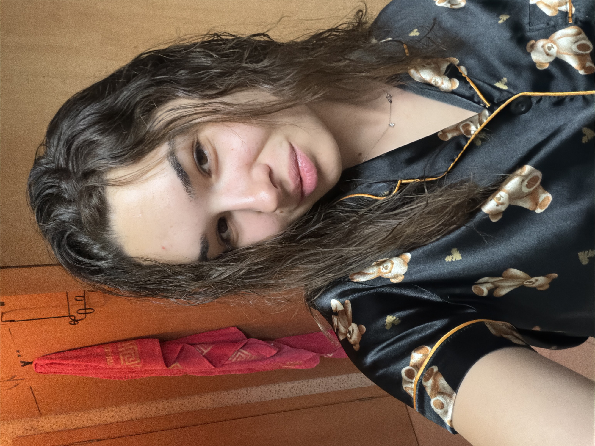 A woman is posing for a selfie in an indoor setting. She has dark, curly hair and is wearing a black pajama top with a teddy bear pattern and yellow trim. She is wearing a delicate necklace. To the left of the woman, there is a red folded towel, hung on a wall with markings. It's hard to determine exact location of the photograph but could be Sofia, Bulgaria.