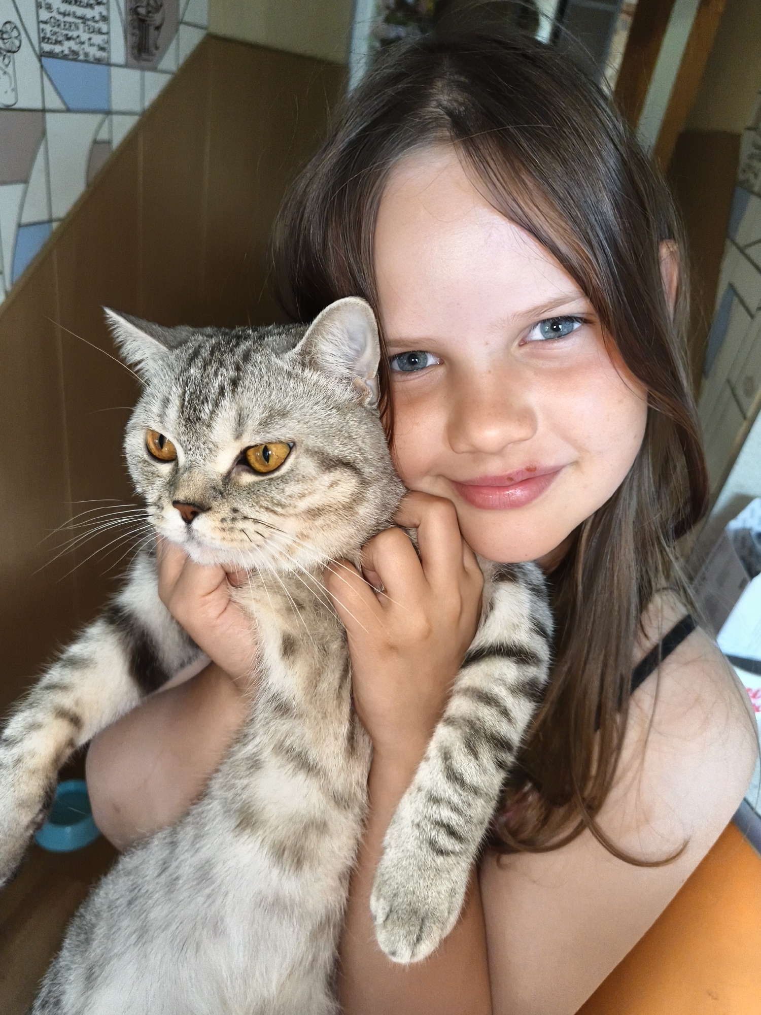 A young girl with long, brown hair and blue eyes is holding a grey tabby cat. The girl is smiling and looking at the camera. The cat is looking to the left with its mouth slightly open. The background is blurry but appears to be indoors with some tiled walls and wooden furniture. The girl is wearing a black tank top. The cat is being held close to the girl's chest.