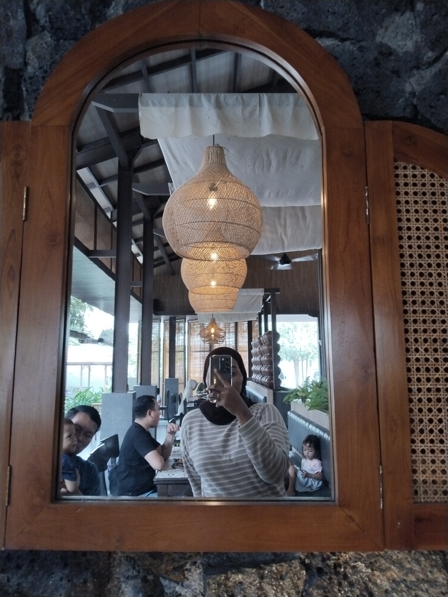 In Kecamatan Pati, Indonesia, a person is taking a selfie in a mirror of what appears to be a restaurant or cafe. The mirror is framed in wood and set into a stone wall. The reflection shows several people, including the photographer holding a smartphone. To the left, a man is holding a child. In the background, another man is seated at a table, and a young child is sitting on a bench. Three large woven lamps hang from the ceiling, reflecting in the mirror. The environment suggests an indoor seating area with natural light filtering in from the outside.