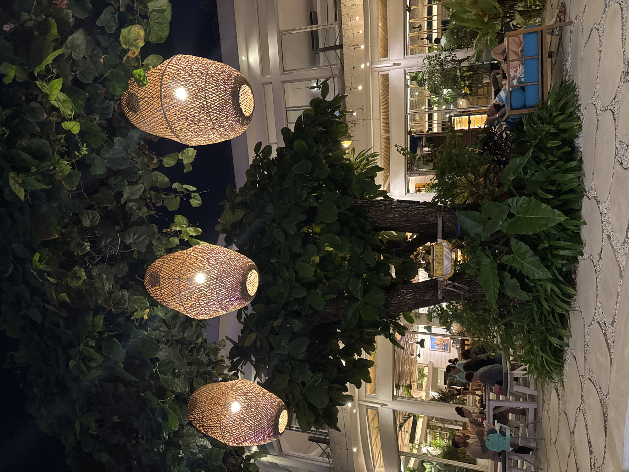 The image shows an outdoor seating area at night, likely a restaurant or cafe, in Kuta, Indonesia. Three large, woven pendant lamps hang from tree branches above. In the background, a two-story building with large windows and balconies is illuminated. People are seated at tables, eating and socializing. One group appears to be a family with a young child. To the right, two individuals are relaxing on a blue outdoor sofa amidst lush greenery. The ground is paved with large, light-colored stones interspersed with gravel. The overall atmosphere is relaxed and tropical, with the warm glow of the lamps and string lights creating an inviting ambiance.