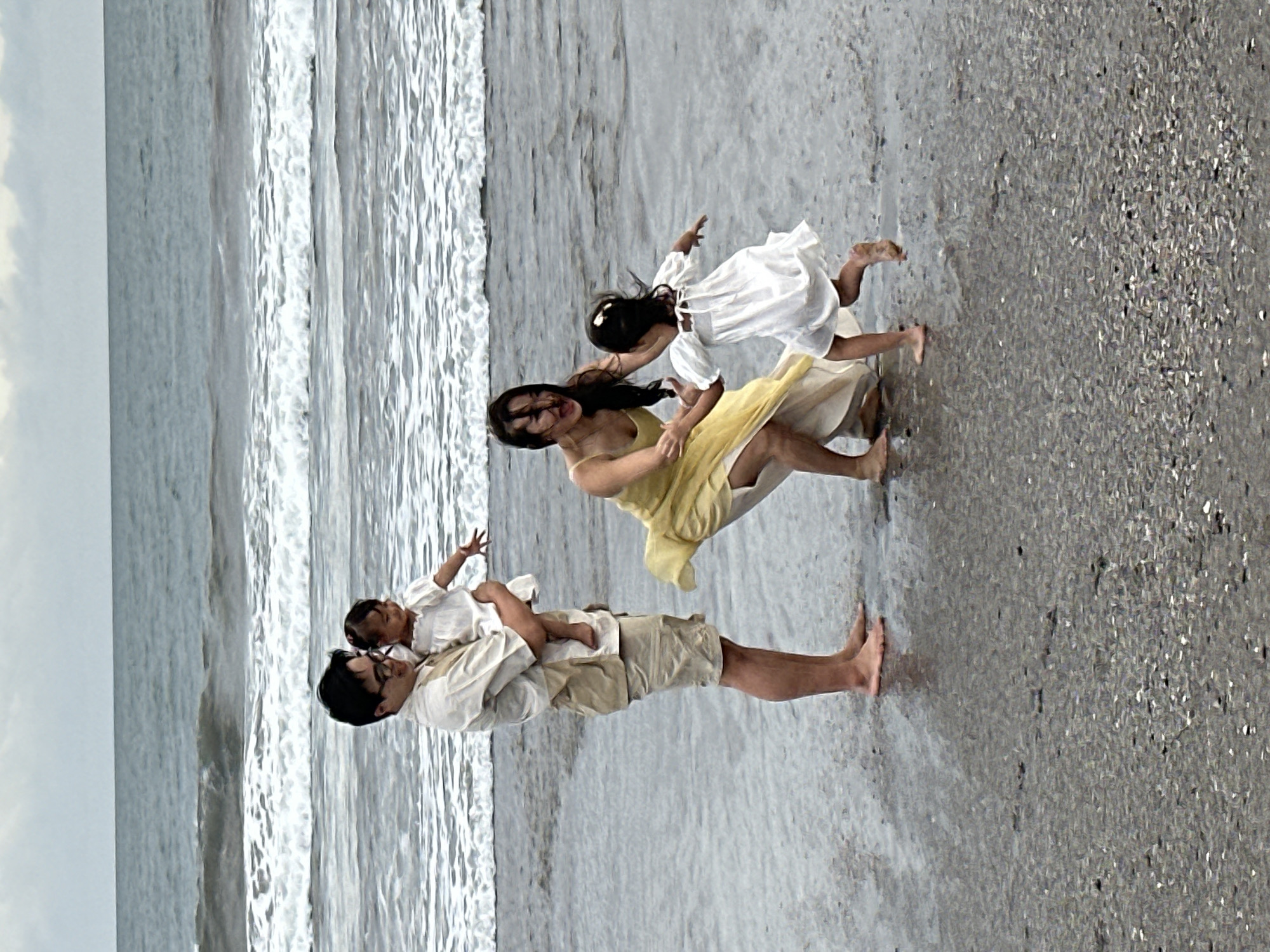 A family of four is standing on a wet, sandy beach in North Kuta, Indonesia. The father, wearing a light-colored shirt and khaki shorts, is holding a baby in his arms. The mother, in a yellow dress, is interacting with a young girl in a white dress who is running away from her. The ocean is in the background with waves breaking on the shore. The sky is overcast.