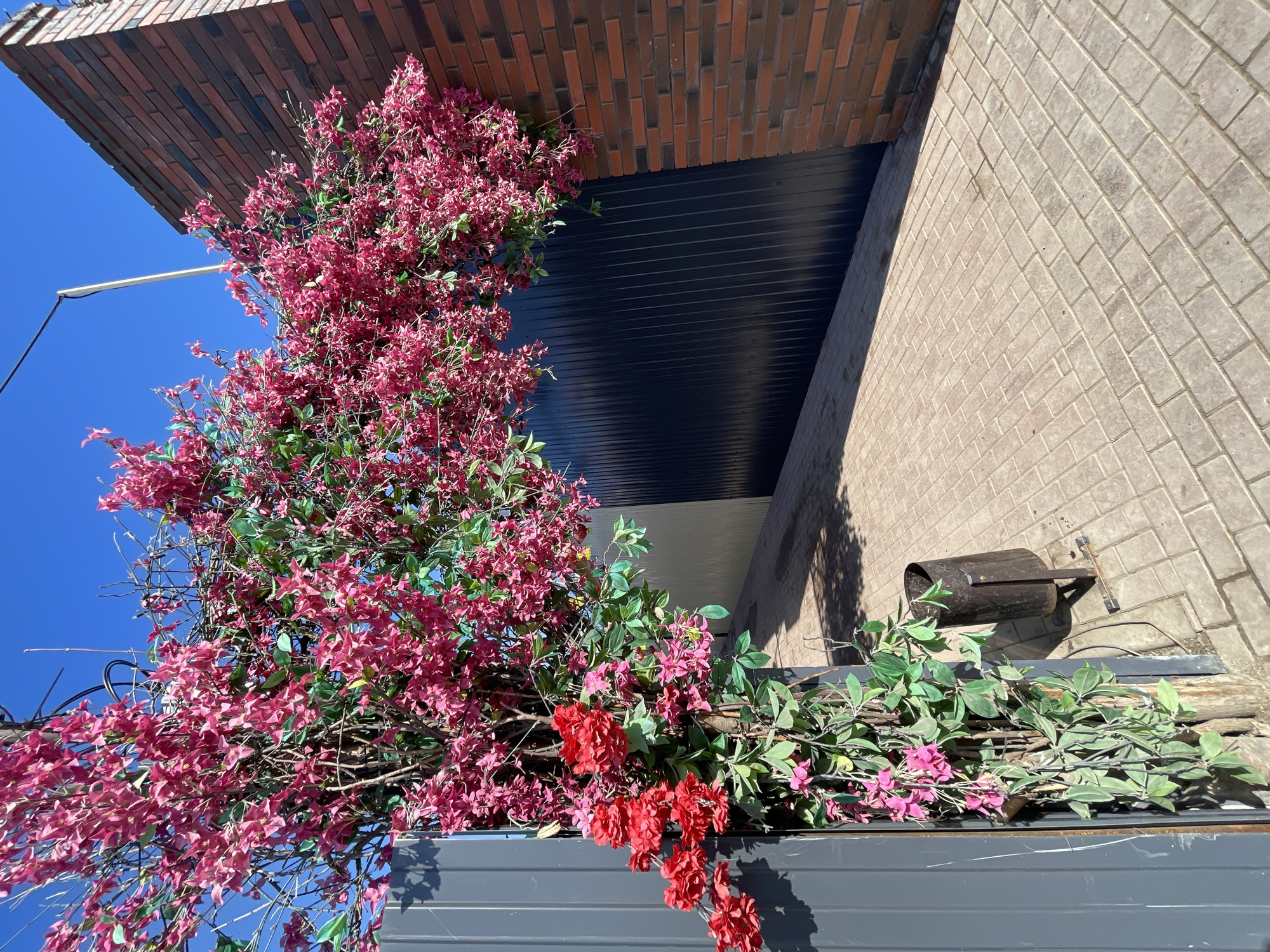 The image depicts a corner of a building with brick and gray tiled exterior walls under a clear blue sky. A section of the building features dark gray horizontal paneling. A large artificial floral arrangement dominates the left side of the frame, featuring pink and red flowers interspersed with green leaves. A small, dark-colored cylindrical object with supporting metal rods is mounted on the tiled wall. The photo is taken from a low angle, tilting upwards towards the building. The location is Osa, Russia.