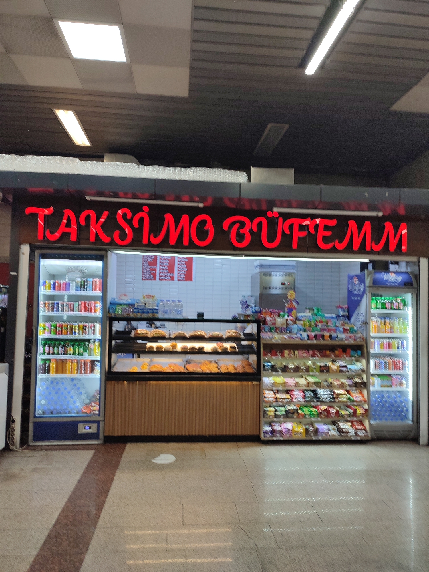 The image shows a storefront with bright red neon lettering that spells out "TAKSİMO BÜFEMM" above a display of beverages and food items. To the left, a refrigerated display case is filled with bottles and cans of drinks. To the right of the beverage case, there is a wooden counter topped with a glass display case containing various pastries and baked goods. Behind the counter, shelves are stocked with packaged snacks and other food items. A second refrigerated display case is visible on the far right, also containing drinks. The floor is tiled with a pattern of light and dark brown and beige stones. The overall environment appears to be an indoor establishment, likely a convenience store or a small eatery, situated in the city of Beyoğlu, Turkey.