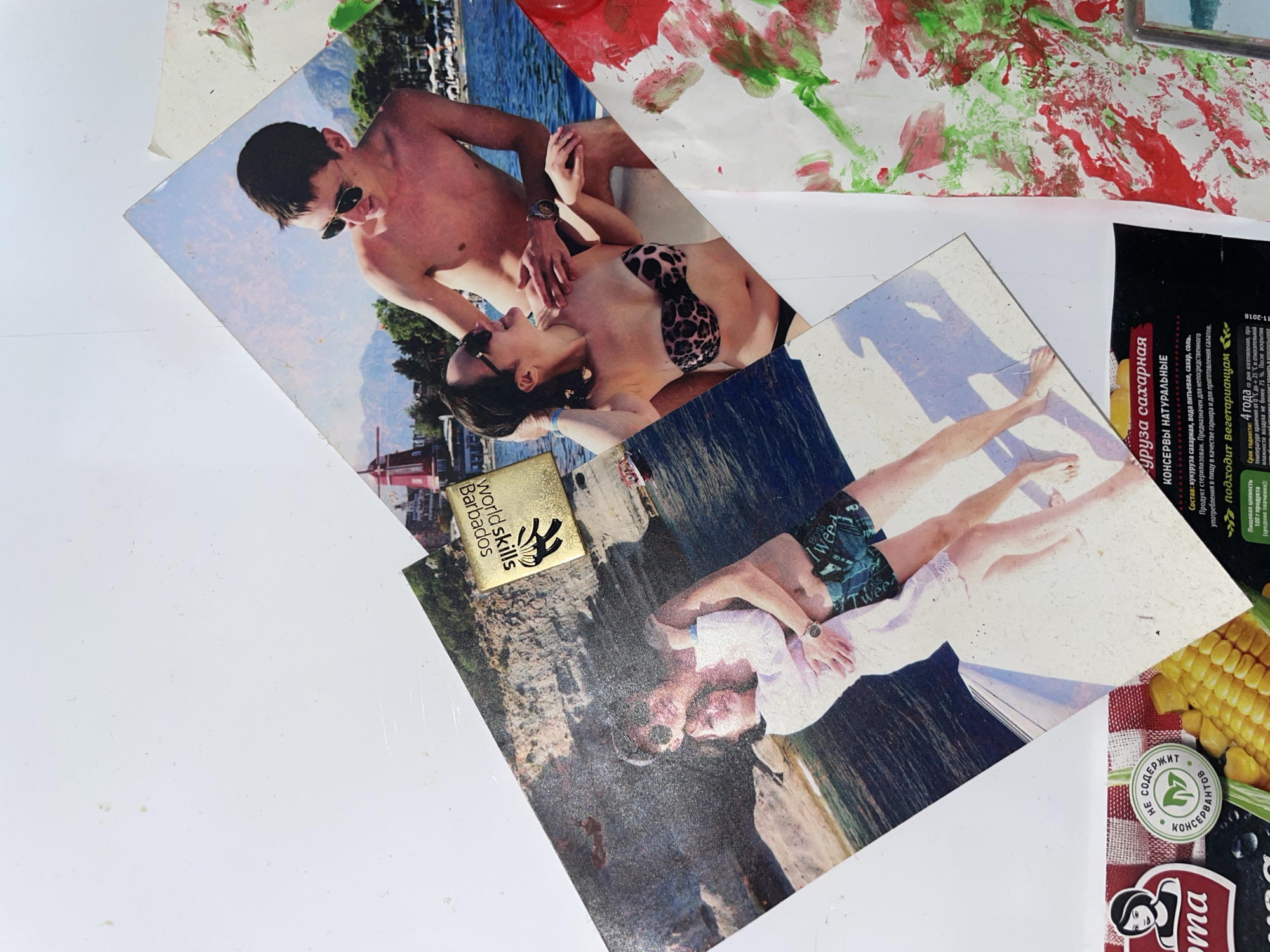 This image displays a collage of photographs and painted elements, predominantly focusing on beach and vacation scenes. At the center, two photographs are layered. The top photograph captures a couple embracing on a boat, with the man wearing patterned swim trunks and the woman in a leopard-print bikini top and sunglasses. Behind them, a younger couple is visible on another boat, with a man in sunglasses and a woman in a bikini, with a body of water and mountainous landscape in the background. A small, gold-colored badge with the text "World Skills Barbados" is attached to the lower left portion of the top photograph.
Below these photographs, a portion of a product label is visible, featuring text in Russian. The visible text includes "НЕ СОДЕРЖИТ КОНСЕРВАНТОВ" (does not contain preservatives) and references to corn. To the right of this label, a section of a painting with red and green abstract brushstrokes is present, suggesting artistic expression. The overall setting appears to be an indoor surface, likely a wall or a table, as evidenced by the flat white background and the way the photographs and painted elements are arranged. There are no discernible elements within the image that specifically indicate the location within Laishevo, Russia.