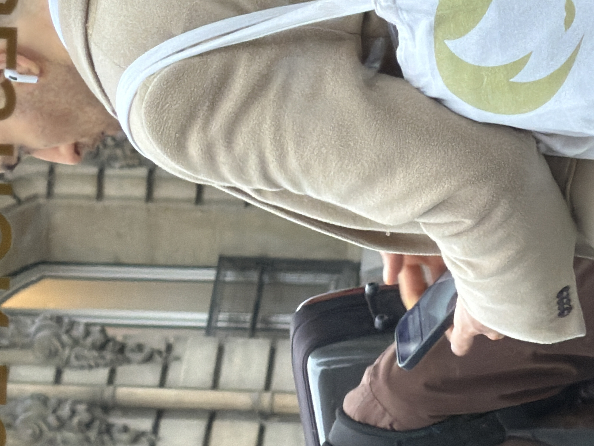A man with glasses and a white earbud is seen from the shoulders up, wearing a tan jacket and a white bag with a yellow symbol. He is holding a dark smartphone. The background shows architectural details suggesting the city of Paris, France, specifically a building facade. The man is pulling a dark grey suitcase.
