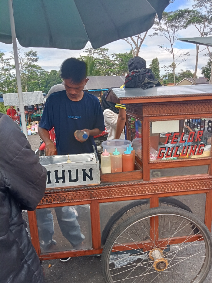 A street vendor is captured in a daylight scene in Singaparna, Indonesia, preparing food from a mobile cart. The main subject is a man wearing a dark blue t-shirt with "Volcom Stone" written on it, focused on his task. He holds a clear plastic bottle in his right hand over a silver metal container labeled "IHHUN" in large, block letters. A skewer with a piece of food is visible within this container, suggesting he is making egg rolls, a popular Indonesian street snack.
The food cart itself is made of intricately carved wood with glass panels. On the upper section, a prominent red sign spells out "TELOR GULUNG" (Egg Rolls). Below this sign, various items like small plastic bags are stored. Next to the cooking area, there are several plastic squeeze bottles containing different sauces, likely for customization of the dish. The cart has large spoked wheels, one of which is visible on the right side.
In the immediate foreground, the left shoulder and arm of a person, possibly a customer, dressed in a dark jacket, partially obscure the view. Behind the vendor, another individual is partially visible, wearing a hijab and a light-colored shirt. The background reveals an outdoor setting with lush green trees under an overcast sky, hinting at a market or public area with other stalls. A large light-colored umbrella shades the vendor and his cart from above. The overall scene depicts a typical bustling street food environment.