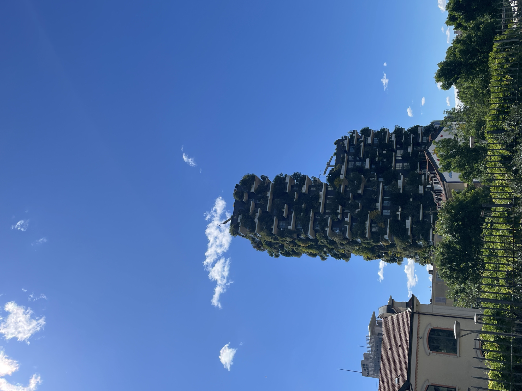 The image shows the Bosco Verticale (Vertical Forest) in Milan, Italy. It is a pair of residential towers with numerous trees and plants growing on their balconies, set against a bright blue sky with scattered white clouds. The surrounding cityscape includes traditional buildings with red-tiled roofs and foliage from other trees and vegetation. A portion of a metal fence is visible in the foreground.