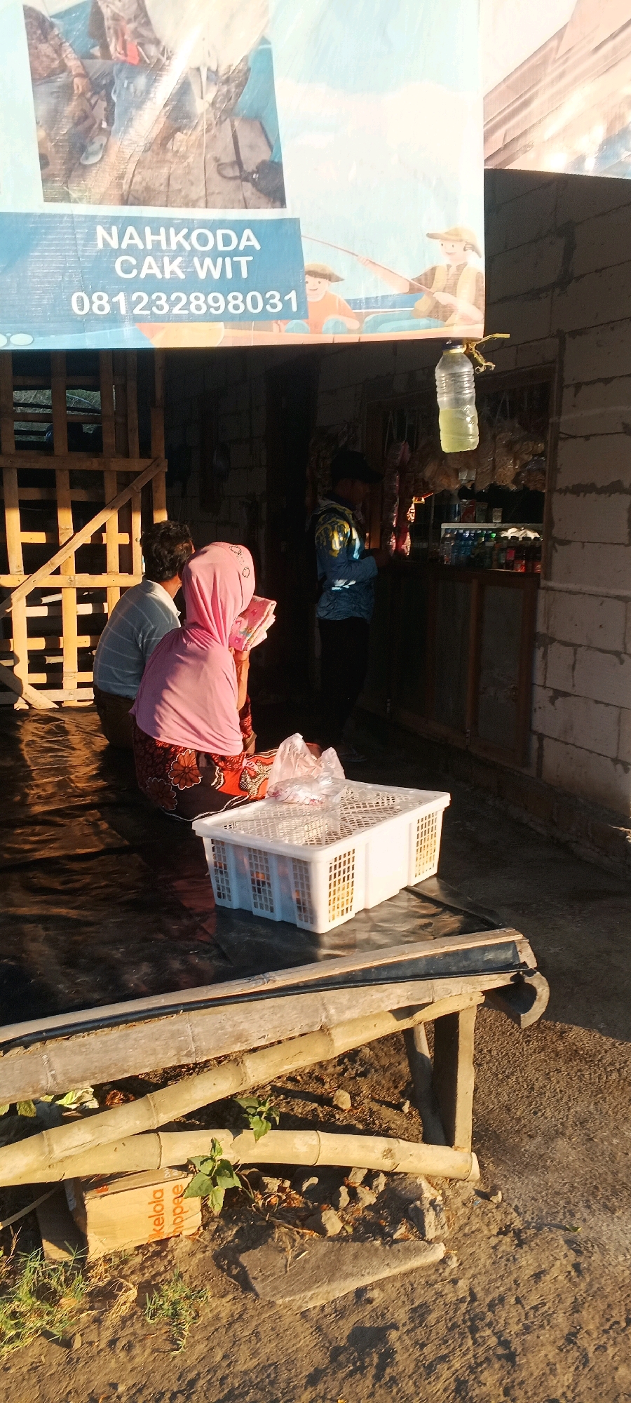 In the city of Tambak Kalisogo, Indonesia, a scene unfolds in front of a modest structure constructed from cinder blocks. The structure appears to be a small shop or stall, with shelves visible inside displaying various bottles and goods. Hanging from a wire above the shop entrance is a plastic water bottle containing a yellow liquid.
In the foreground, two individuals are seated on a dark-colored tarp spread on the ground. One person, a man wearing a striped long-sleeved shirt, is seen from the back. Beside him, a woman wearing a pink hijab and a patterned skirt is also seated, holding a book. Between them and slightly to the right, a white plastic crate is placed on the tarp. The crate contains items covered in plastic bags, possibly groceries or merchandise.
To the left of the seated individuals, a wooden structure made of interconnected planks is visible, likely part of the stall's framework or storage. Further back and to the right of the shop entrance, another person, a man wearing a blue patterned shirt and a black cap, stands near the shop counter, facing away from the camera and possibly interacting with the shopkeeper or examining goods.
The ground around the structure is dirt, with scattered debris including rocks and dried vegetation. Several bamboo poles are arranged on the ground in the lower section of the image, and a small cardboard box with Indonesian writing is also present among the bamboo. The overall lighting suggests it is either early morning or late afternoon, with strong shadows indicating the direction of the sun. A banner with the text "NAHKODA CAK WIT" and a phone number "081232898031" is prominently displayed in the upper left portion of the image, depicting illustrations of people fishing.