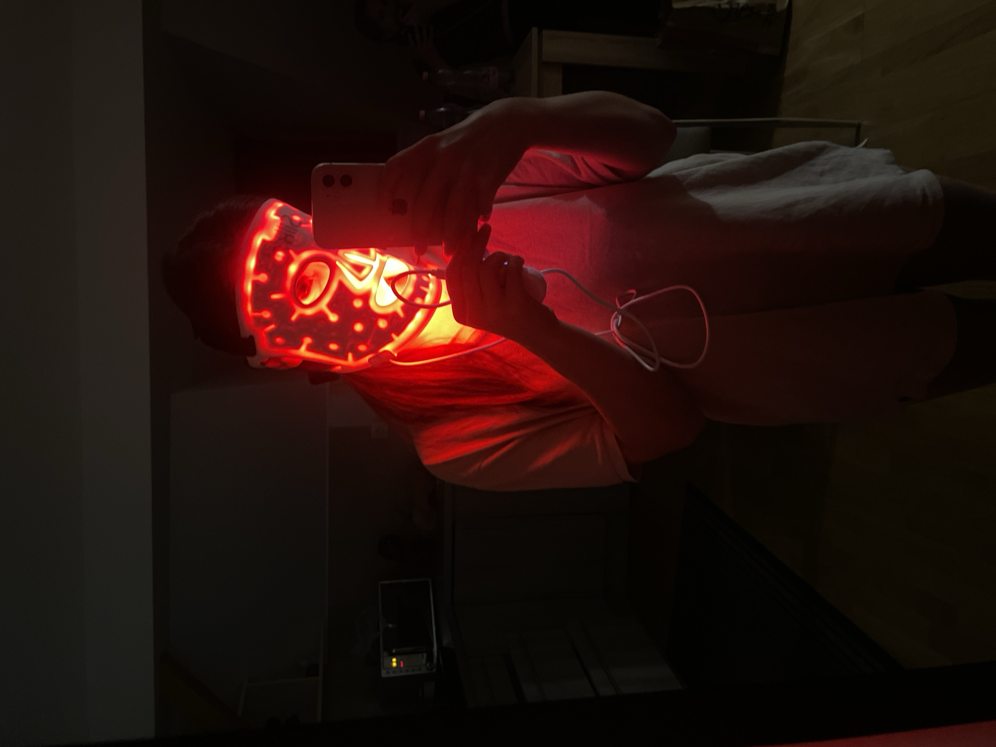 A person is wearing a red light therapy mask and holding a cell phone in a dimly lit room. The mask emits a bright red light, illuminating the person's face and hands. The person is wearing a light-colored shirt. The background is dark, with vague shapes of furniture visible. The location is not identifiable as being in the city of Hévíz, Hungary from the image alone.