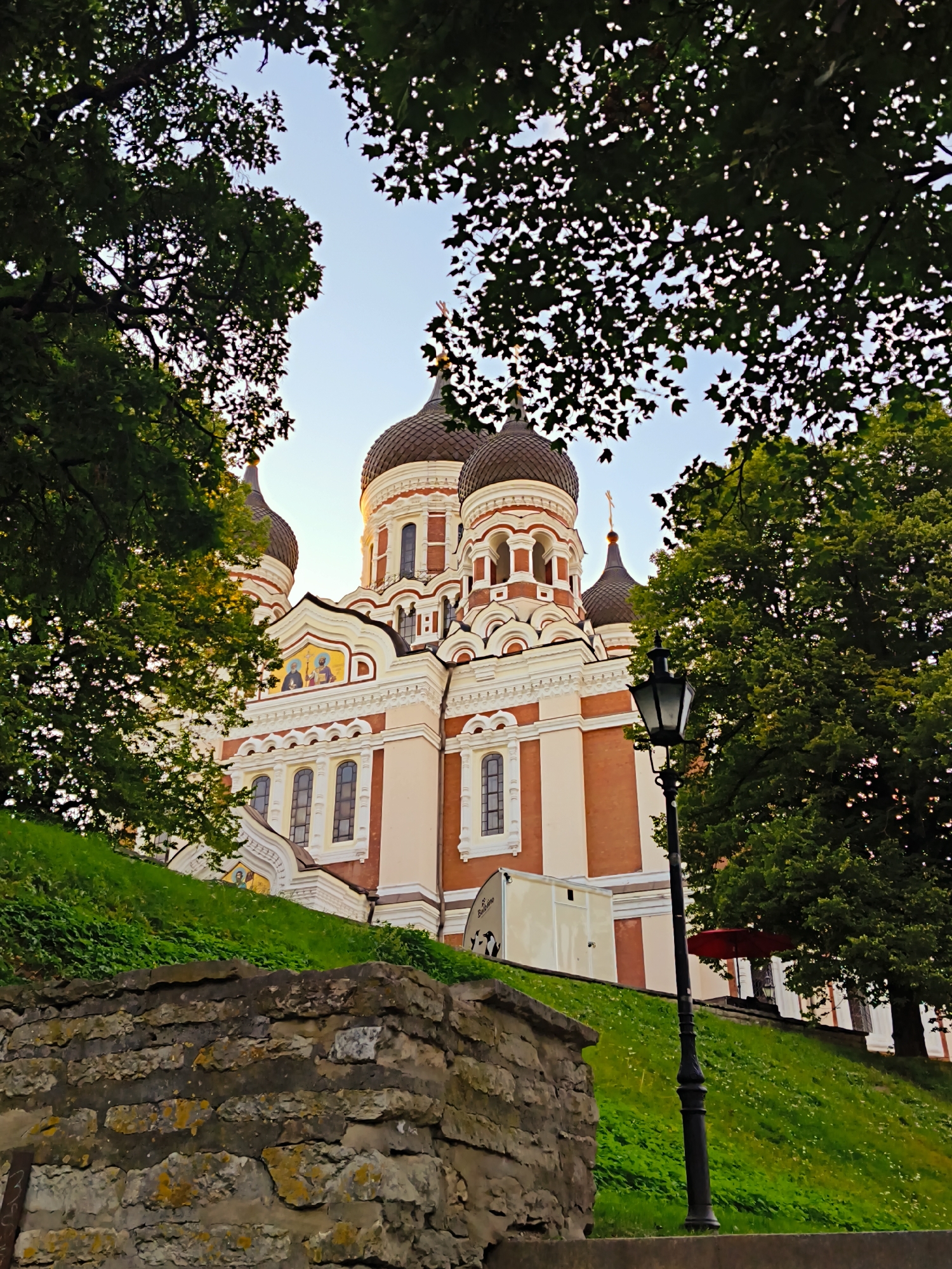 The image shows the Alexander Nevsky Cathedral, a prominent Orthodox church located in Tallinn, Estonia. The cathedral, with its distinctive onion domes and red and white facade, is situated on a grassy hillside. The foreground features a stone wall and a vintage lamppost, while the background is dominated by lush green trees. The sky above is a clear, soft blue, suggesting it might be late afternoon or early evening. There are no people visible in the image.