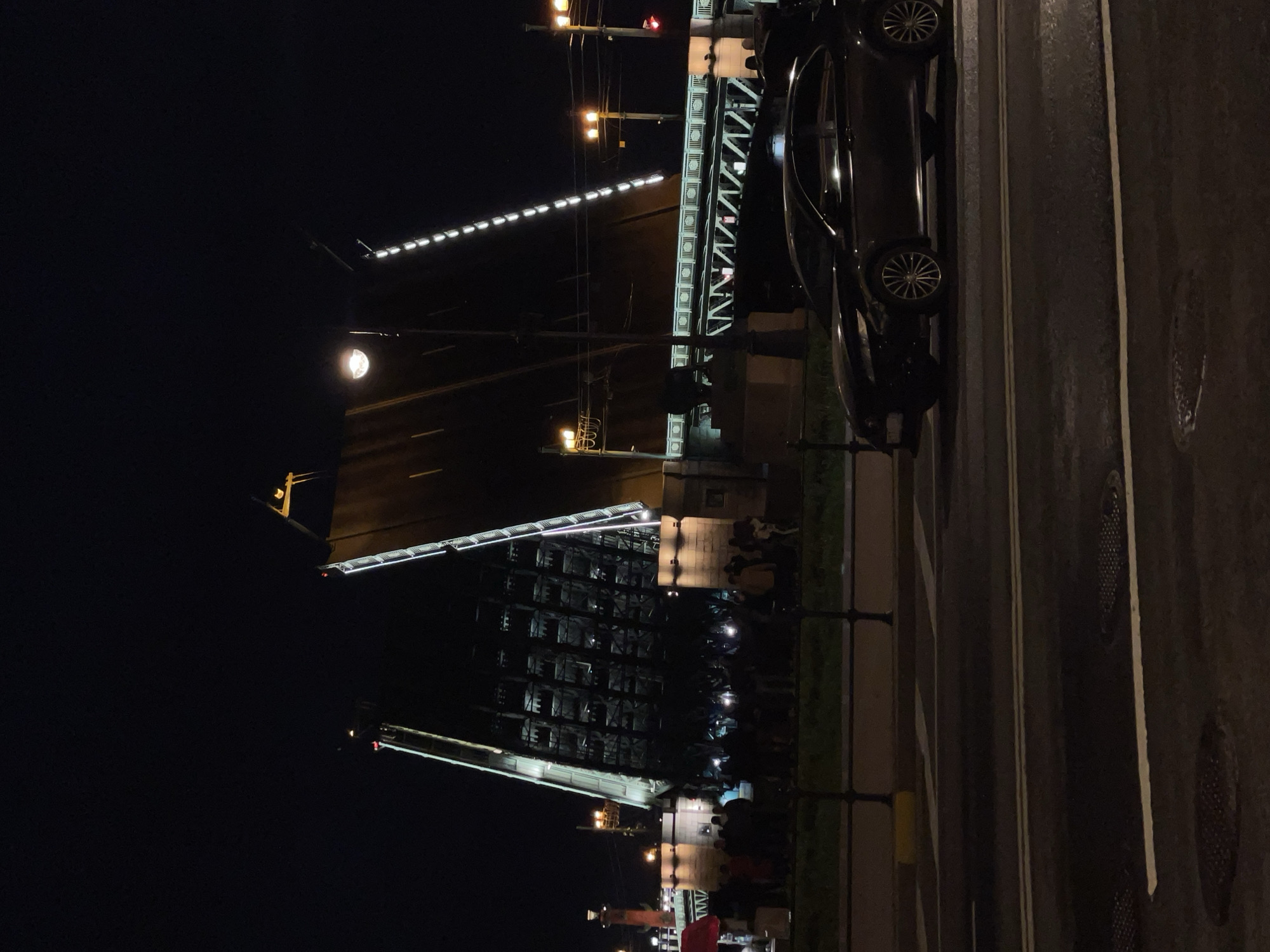 At night, a bridge in Saint Petersburg, Russia, is raised, illuminated by multiple rows of lights. A dark-colored car is parked on the road next to a black metal fence, with spectators gathered to watch the bridge opening.
