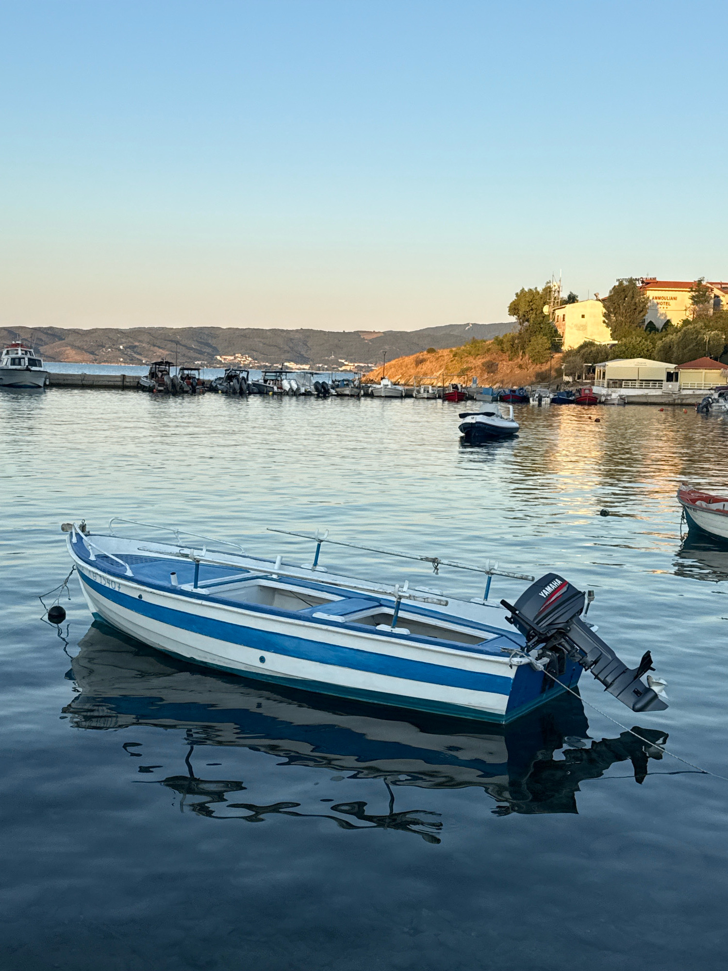 The image depicts a serene harbor scene in Ammouliani, Greece, during what appears to be early morning or late afternoon given the soft light and clear sky. In the foreground, a small, white and blue motorboat is moored, its reflection shimmering on the calm water. The boat is equipped with a Yamaha outboard motor. In the middle ground, several other boats are docked along a pier, with a few larger vessels visible further out. The background features a gently sloping, tree-covered hill with buildings situated on its crest, including a prominent yellow building labeled "AMMOULIANI HOTEL." The overall atmosphere is peaceful and idyllic, characteristic of a Greek coastal village. There are no people visible in the image.