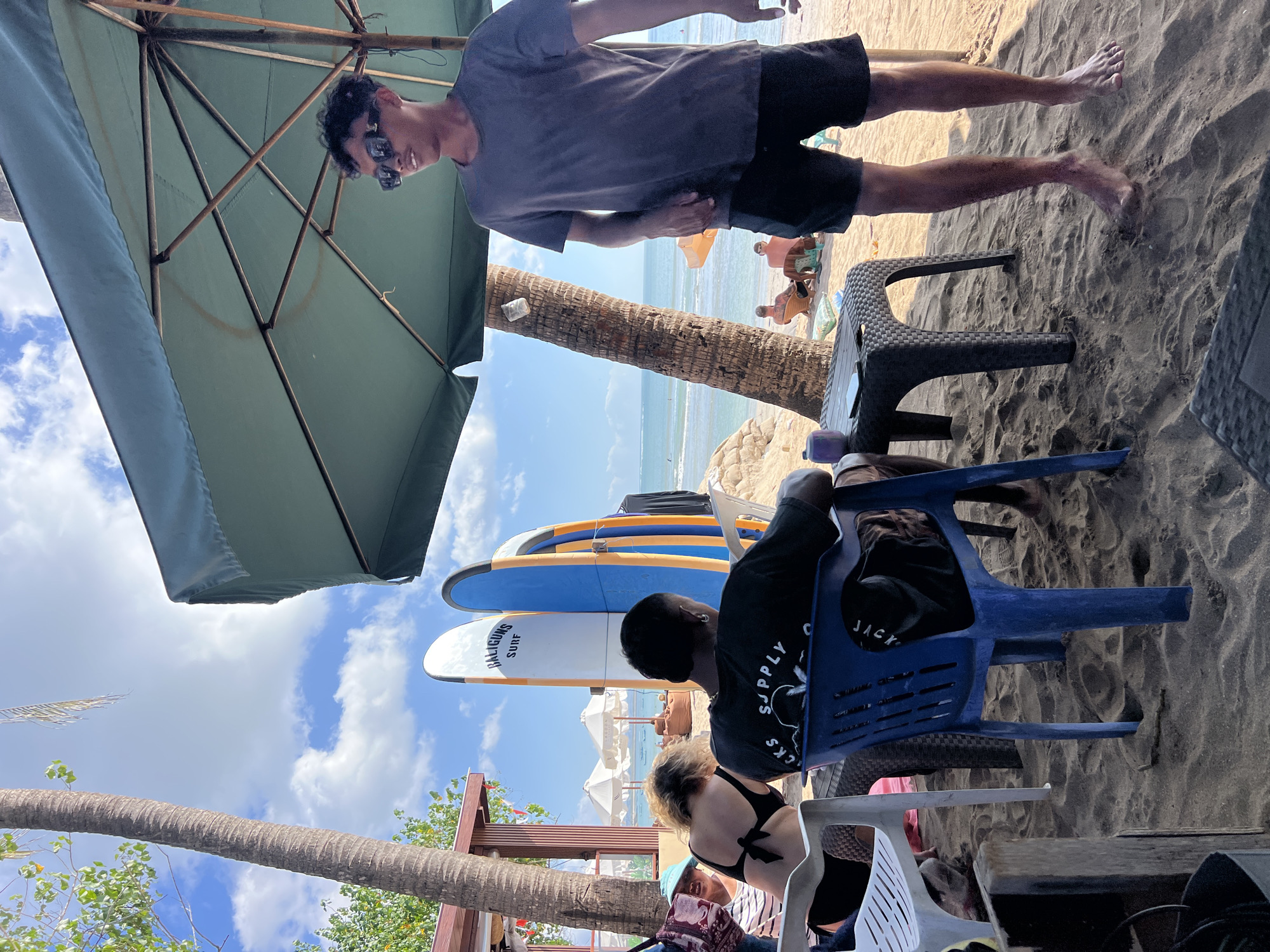A sunny beach scene in Kuta, Indonesia, features several individuals relaxing at a beachfront establishment. In the foreground, a young man wearing sunglasses and a dark grey t-shirt stands near a large green umbrella, facing right. To his left, a person in a blue plastic chair faces away from the camera, wearing a black t-shirt with white text that reads "JACK SUPPLY CO." Next to him, a woman with blonde hair, wearing a black bikini top and fishnet stockings, is seated on a white chair and looking towards the left. Behind them, three surfboards are propped up vertically: a white board with "BALIGUNS SURF" written on it, a blue and orange board, and another blue board. The background shows the ocean with small waves and a clear sky with scattered clouds. Further back, two individuals are seen reclining on beach chairs. The ground is covered in sand, with a few plastic tables and chairs scattered around. Palm trees frame the left side of the image, and a wooden structure appears to be part of the beachfront establishment.