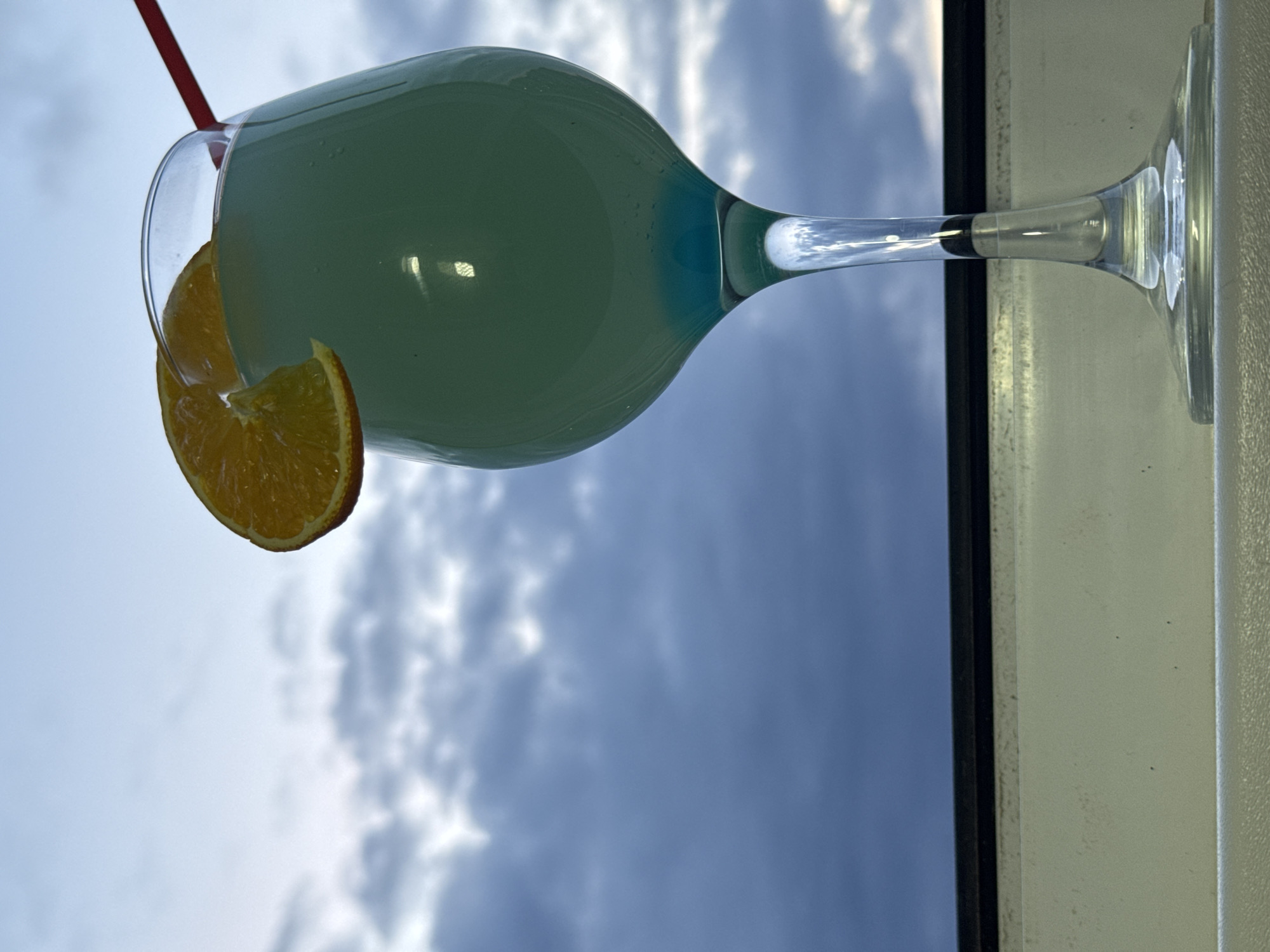 A cocktail in a large glass sits on a windowsill against a backdrop of a cloudy sky. A slice of orange garnishes the rim of the glass, and a red straw is visible. The glass is filled with a light blue or green liquid. The setting appears to be indoors looking outward. There is no indication of this being in Chaikovsky, Russia.