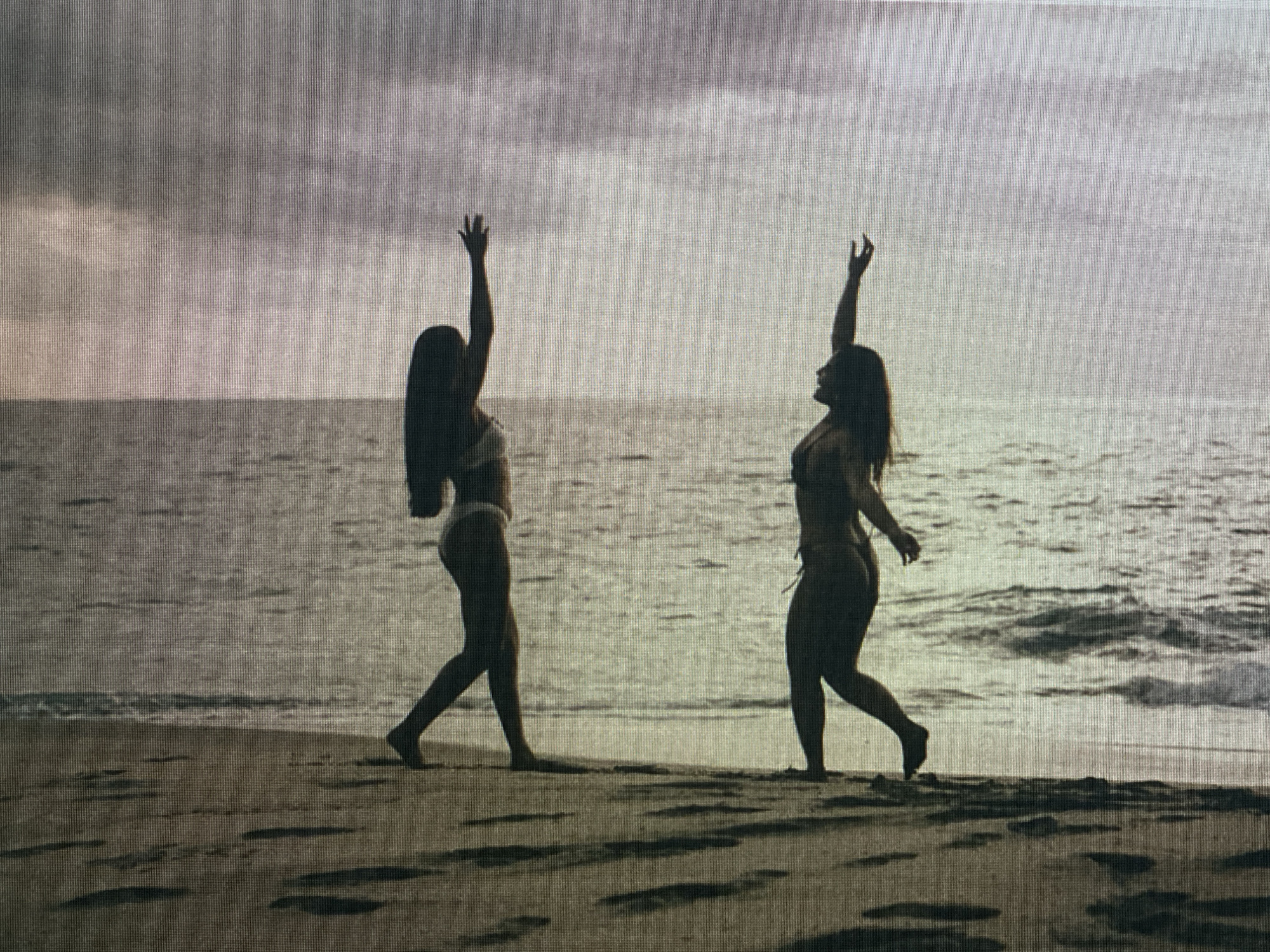 Two women in bikinis are silhouetted against a cloudy sky on a sandy beach. The woman on the left has her left arm raised with her hand open, and her right arm is down by her side. She is facing left. The woman on the right has both arms raised with her hands open, and she is facing right. Both women appear to be dancing or celebrating. The ocean is visible behind them with gentle waves lapping at the shore. The sand on the beach has footprints. The lighting suggests it is either early morning or late evening.