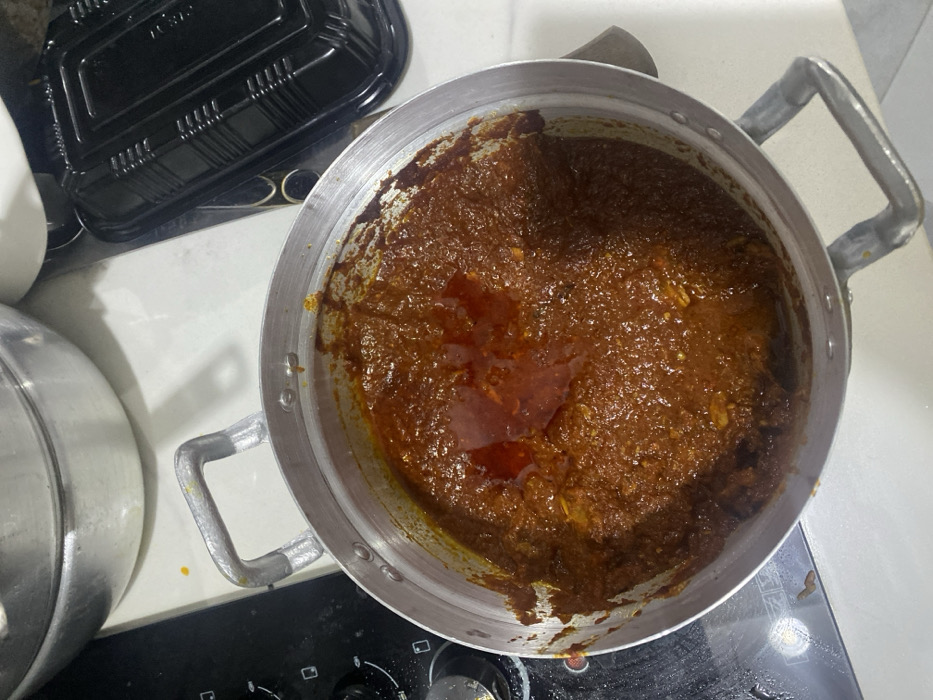 The image displays an overhead view of a large, silver-colored metal pot filled with a thick, dark reddish-brown sauce or stew. The sauce has an oily sheen on its surface, suggesting it is either actively cooking or freshly prepared. Small, lighter-colored chunks, likely pieces of ingredients such as meat, fish, or vegetables, are visible within the rich mixture. The pot is equipped with sturdy, U-shaped metal handles on its sides.
The cooking vessel is placed on a white countertop or stovetop. A black electric or gas hob, with visible control knobs, occupies the lower part of the frame. To the upper left of the main pot, a black plastic takeaway container or its lid is partially visible. Another silver metal pot or lid is partially seen on the far left, further indicating a kitchen setting. The appearance of the dish is consistent with traditional West African cuisine, such as a hearty stew or soup, aligning with the provided location context of Adenta Municipality, Ghana. No people are present in the image, and there is no visible text. The time of day and weather cannot be determined.