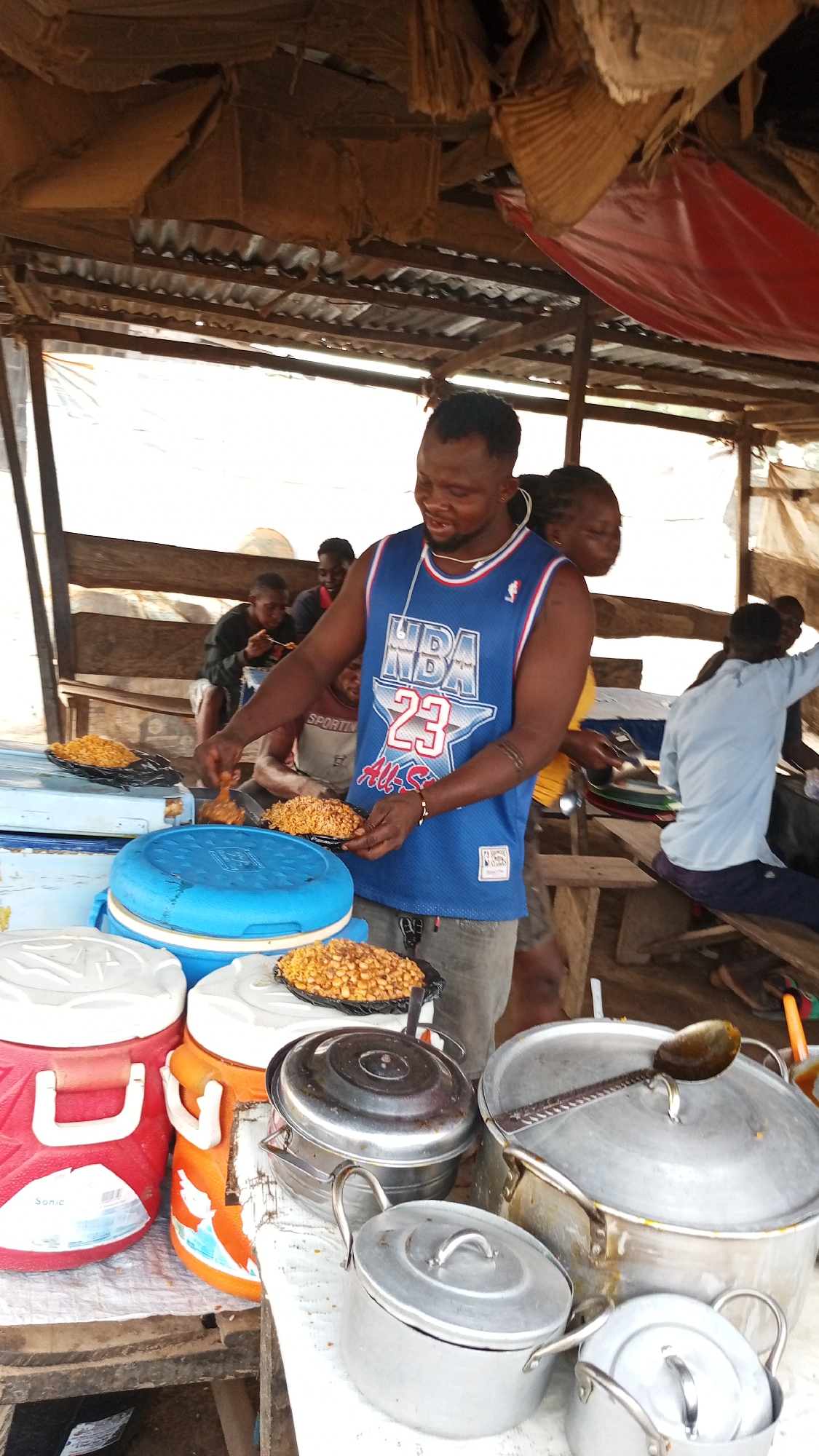 This media file depicts a bustling outdoor street food stall, likely in Ajaokuta, Nigeria. The central figure is a smiling man wearing a blue NBA tank top with "23" and "All-S" visible on it. He is actively serving or preparing food, holding a black plate filled with a yellow, granular dish (possibly a rice or bean dish) and adding what appears to be a piece of fried chicken to it. Another identical plate of food rests on a blue cooler next to him, and a third is visible on a white surface.
The stall is set up under a makeshift shelter with a corrugated metal roof and draped fabrics. On a wooden table, various cooking and serving items are arranged, including several large aluminum pots with lids, one holding a serving spoon, and multiple colorful cooler boxes (red, orange, blue, white). The red cooler has "Sonic" partially visible on its side.
In the background, several other people are present, some seated at wooden benches, appearing to be customers enjoying meals. One person's shirt has the word "SPORTS" visible. The scene conveys a lively, communal atmosphere typical of local food markets, with the vendor's pleasant demeanor suggesting a friendly service. The time of day appears to be daytime, with ample ambient light.