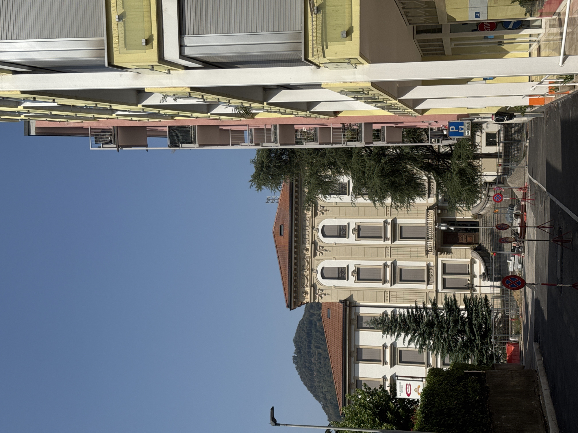 In Chiasso, Switzerland, on a bright, clear day, a street scene unfolds with several buildings and a distant hillside. To the right, a tall, modern building with balconies painted in pink and yellow accents dominates the foreground. Its facade features rows of windows with closed, grey roller shutters and yellow glass railings on some of the balconies. White vertical pillars support the structure.
To the left, a more traditional, cream-colored building with ornate architectural details stands. It has a red-tiled roof and arched windows with dark shutters. Adjacent to this building, a tall, dense evergreen tree is prominently featured. Further left, a hedgerow lines the street.
In the middle ground, construction appears to be underway, with fencing and piles of material visible. Several temporary traffic signs, including one with a red circle and a diagonal red bar over a blue "P" indicating no parking, are placed on the asphalt. A blue parking sign with a coffee cup symbol and "60 MINUTI" is also visible on the right side of the street. In the distance, a lush green hillside rises under the clear blue sky. The street itself is paved and appears empty of people or vehicles.
