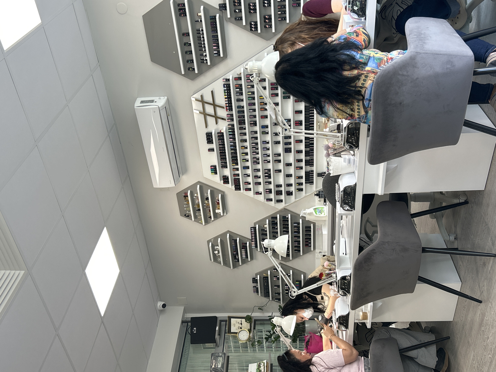 The image shows the interior of a nail salon, likely in Unknown, Unknown. Three women are visible, apparently receiving manicures at three separate stations. The stations have white countertops with overhead adjustable lamps. Behind the manicure stations, the walls are decorated with geometric shelves displaying numerous bottles of nail polish. An air conditioning unit is mounted on the wall. The setting appears clean and professional.