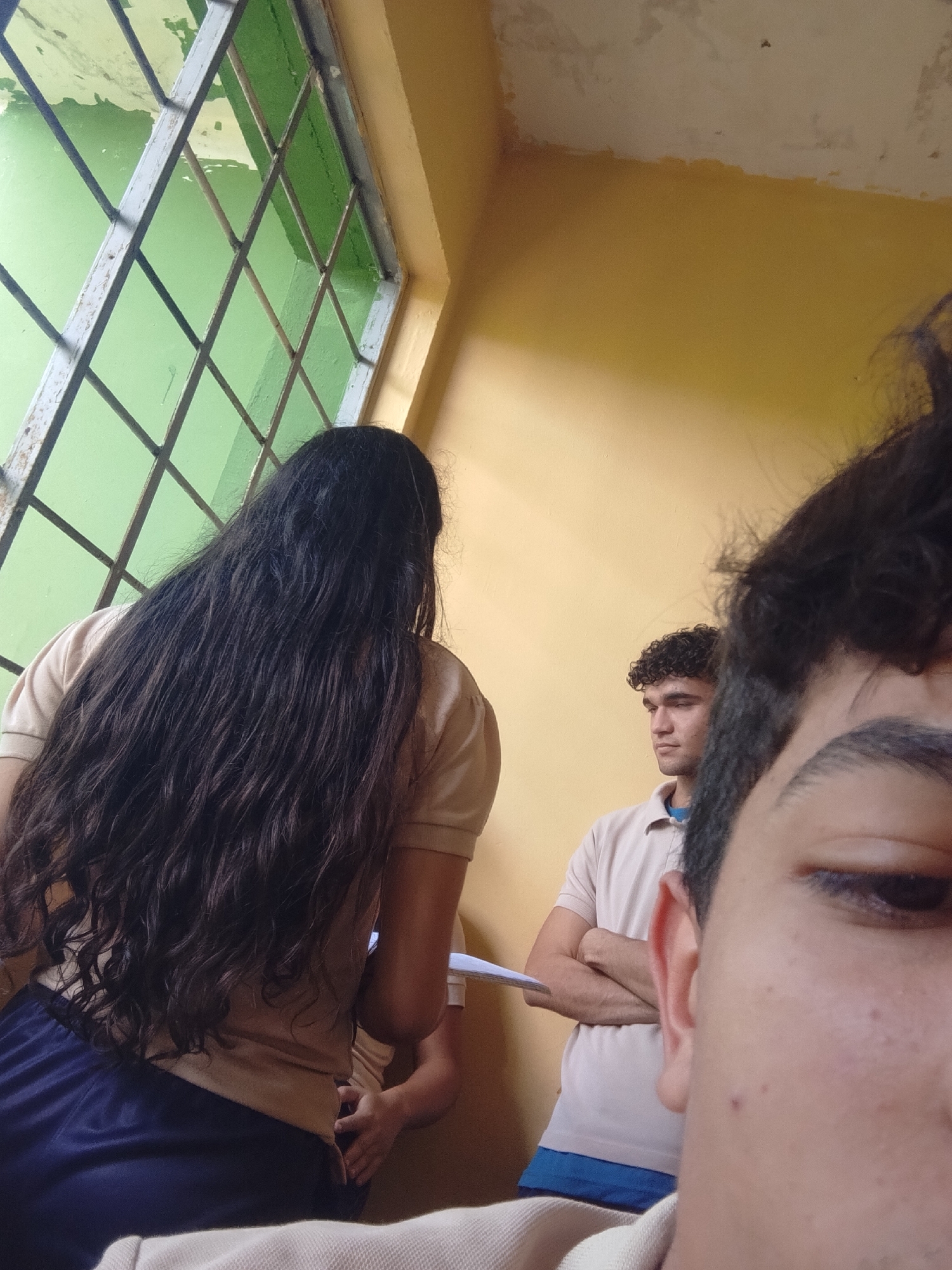 In Santa Ana, Venezuela, an indoor scene depicts at least three individuals near a window with green-painted bars. A person with very long, dark, wavy hair is positioned in the foreground, facing away from the camera and towards the window. This individual is wearing a light-colored short-sleeved shirt and dark blue pants. To the right and slightly further back, another individual with curly dark hair and a light-colored collared shirt stands with their arms crossed, looking to the left. A third person's face is visible in the extreme foreground, partially obscuring the view. The background wall is painted a bright yellow. The lighting suggests daylight entering through the window.