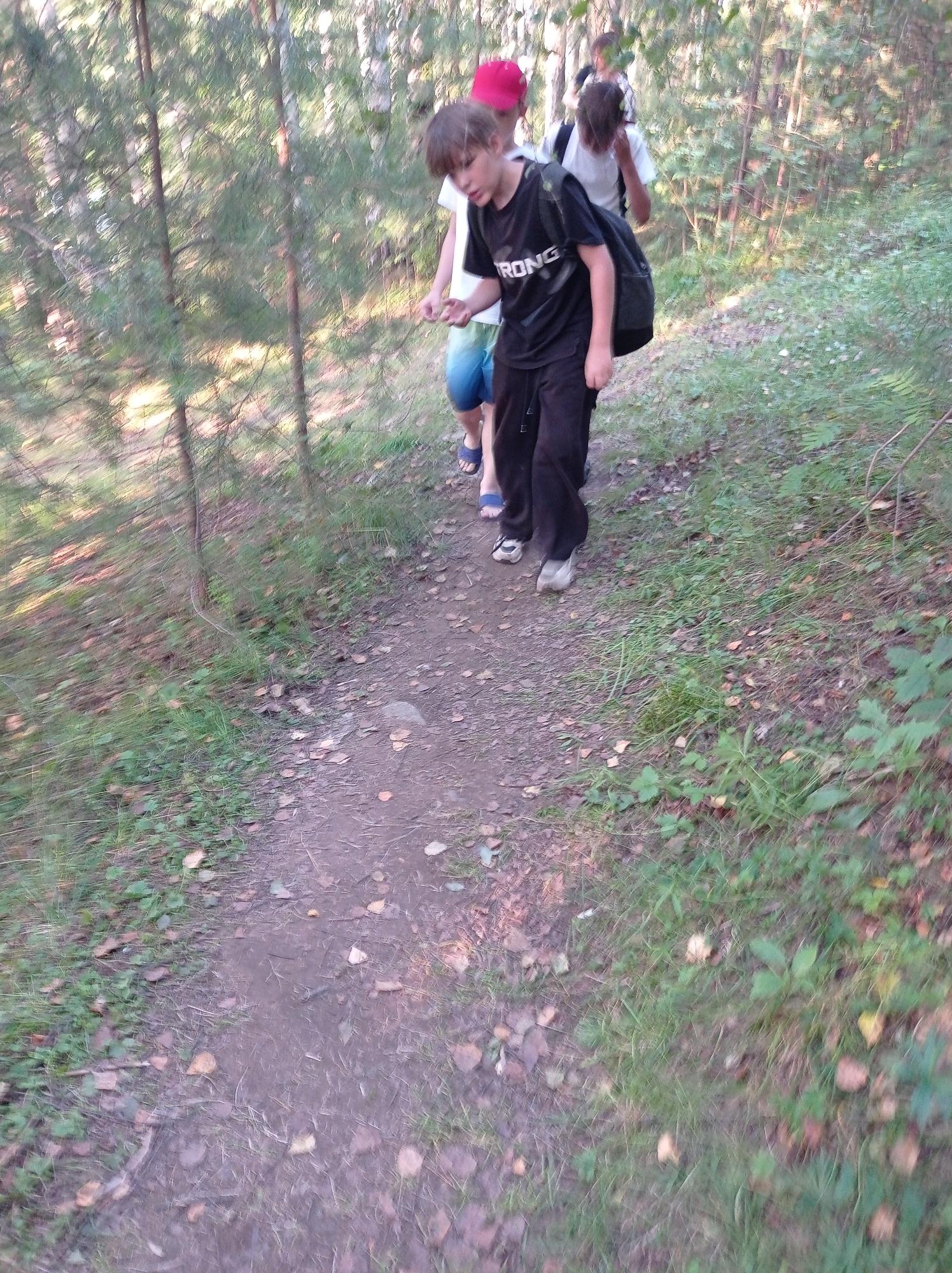 In an outdoor forest setting, several individuals are walking along a dirt path. The foreground shows the legs of a person wearing dark pants and black shoes, their feet on the path. To the left, another person's legs are visible, wearing blue sandals and blue shorts. Behind them, a person in a black t-shirt with the word "RONG" printed on it in white, dark pants, and carrying a black backpack walks on the path. Further back, another person with a red cap is partially visible, and behind them, a person in a white t-shirt and dark shorts is walking, also with a backpack. The path is bordered by vegetation and trees, with fallen leaves on the ground. The lighting suggests it is daytime, with sunlight filtering through the trees. The location is identified as Degtyarsk, Russia.